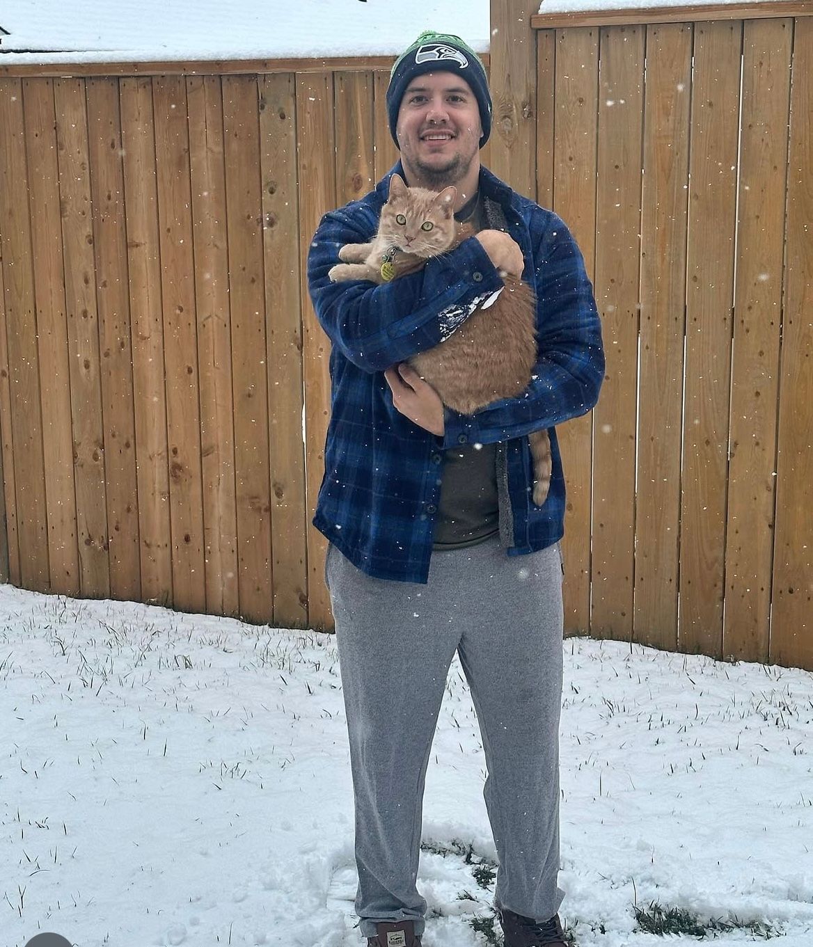 Man holding orange cat in the snow; both bundled in warm clothing near a wooden fence.
