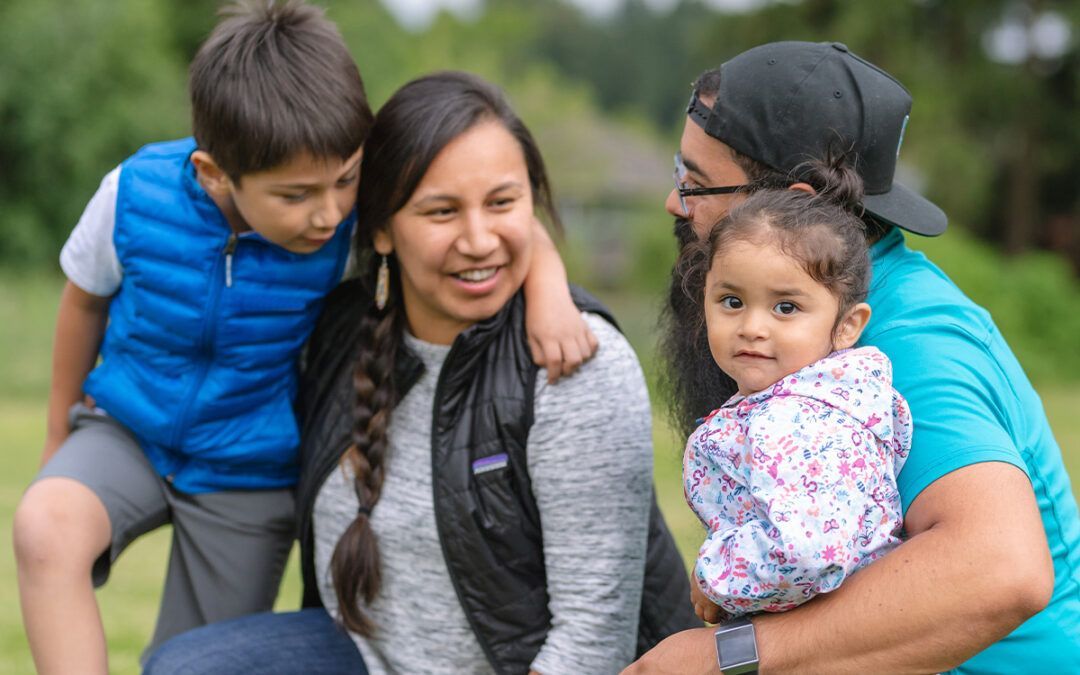Family of four outdoors; a parent holds a toddler, while another parent and child look on.