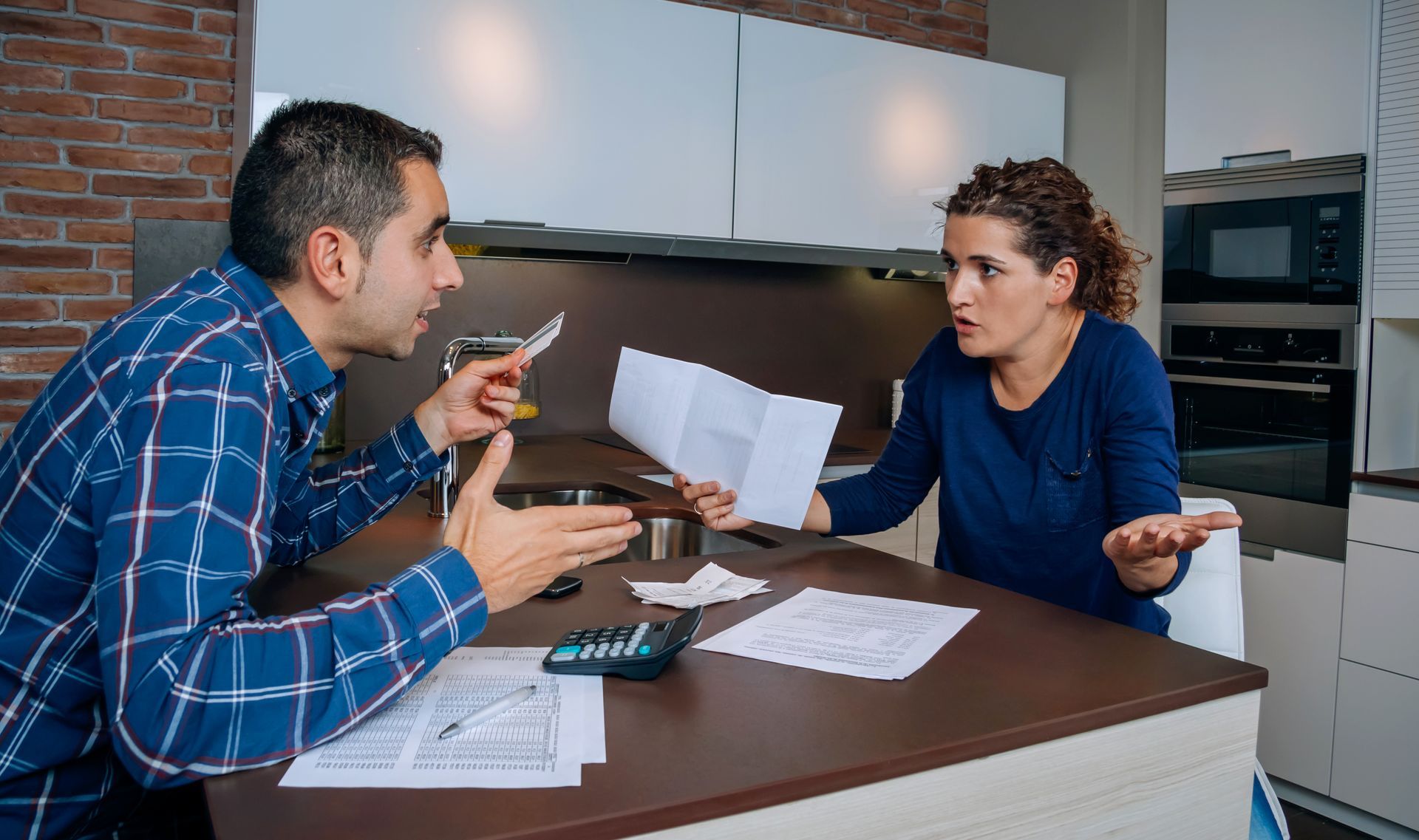 Couple arguing over bills at a kitchen table. Papers, calculator, and frustrated expressions.