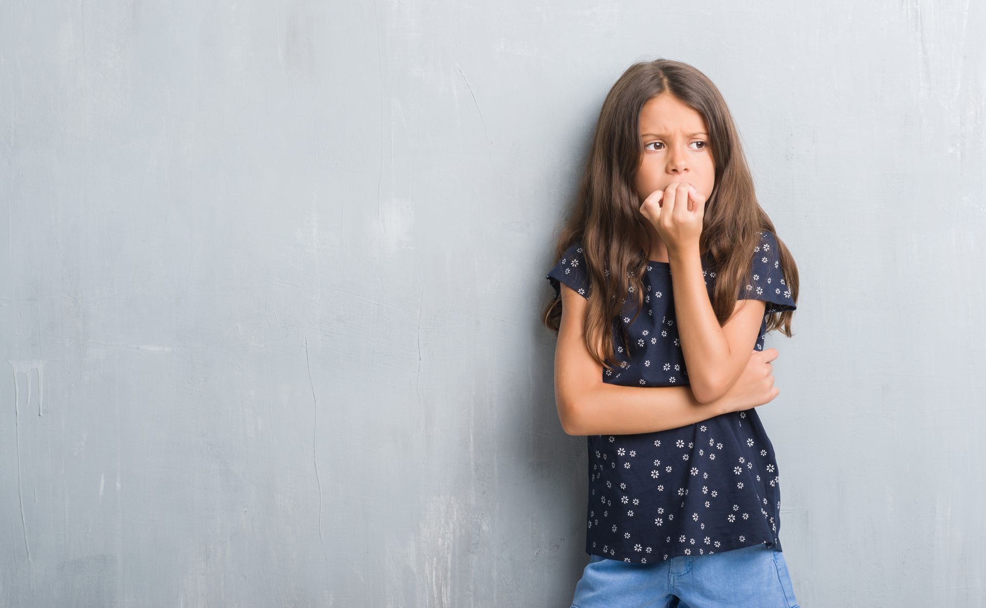 Girl biting nails, looking anxious, arms crossed, standing against a gray wall.