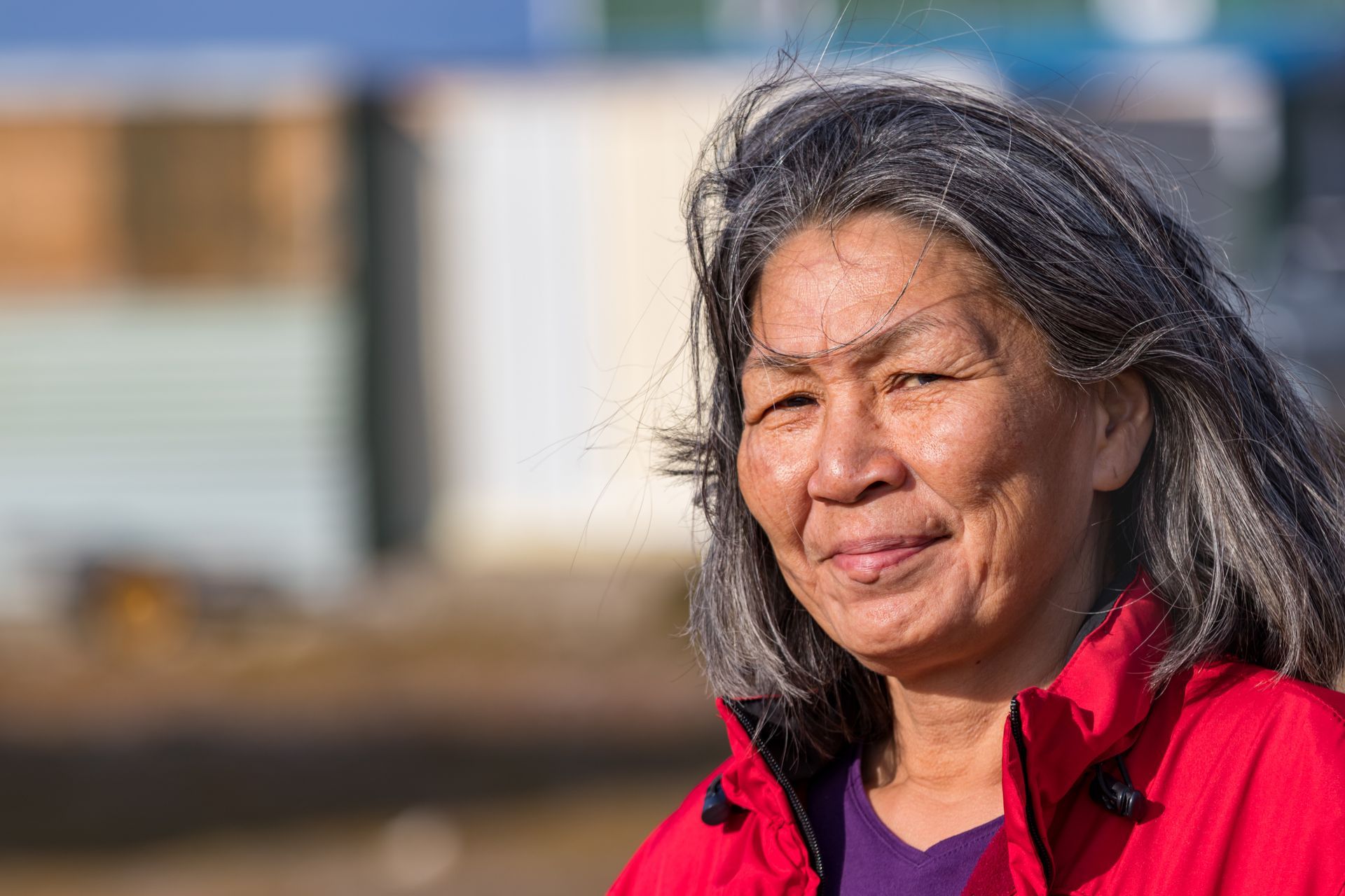 Woman with gray hair smiles, wearing a red jacket, set outdoors in front of buildings.