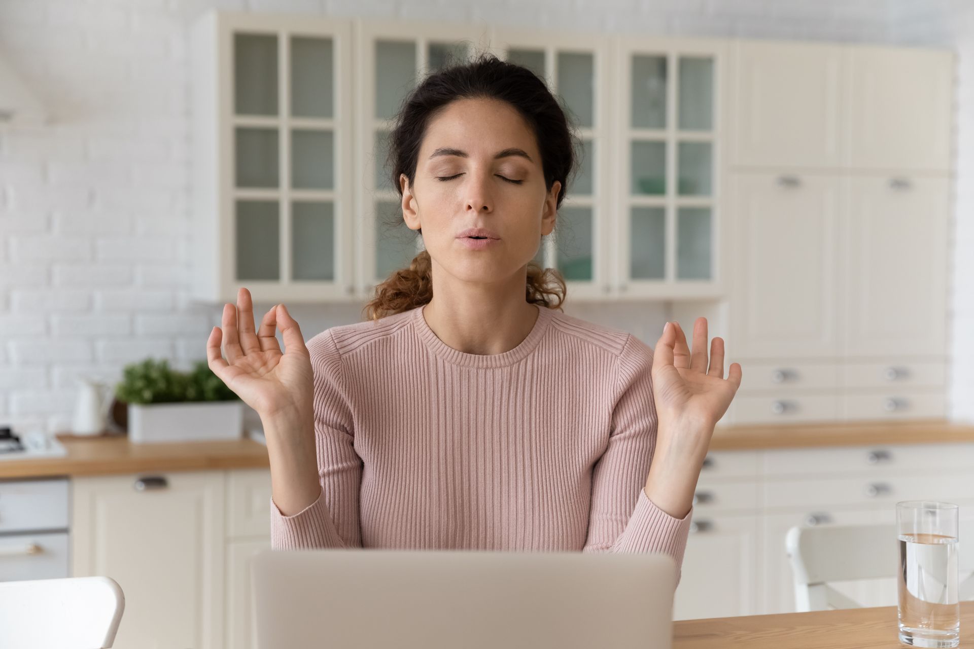Woman meditating at desk with laptop, eyes closed, hands raised, kitchen background.