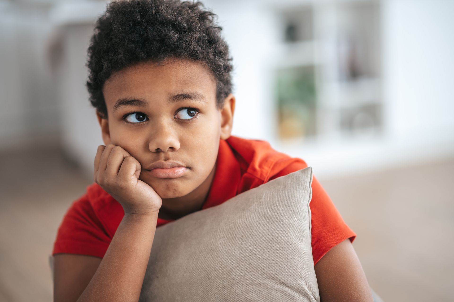 Boy with curly hair, red shirt, and a pout, resting head on a pillow.