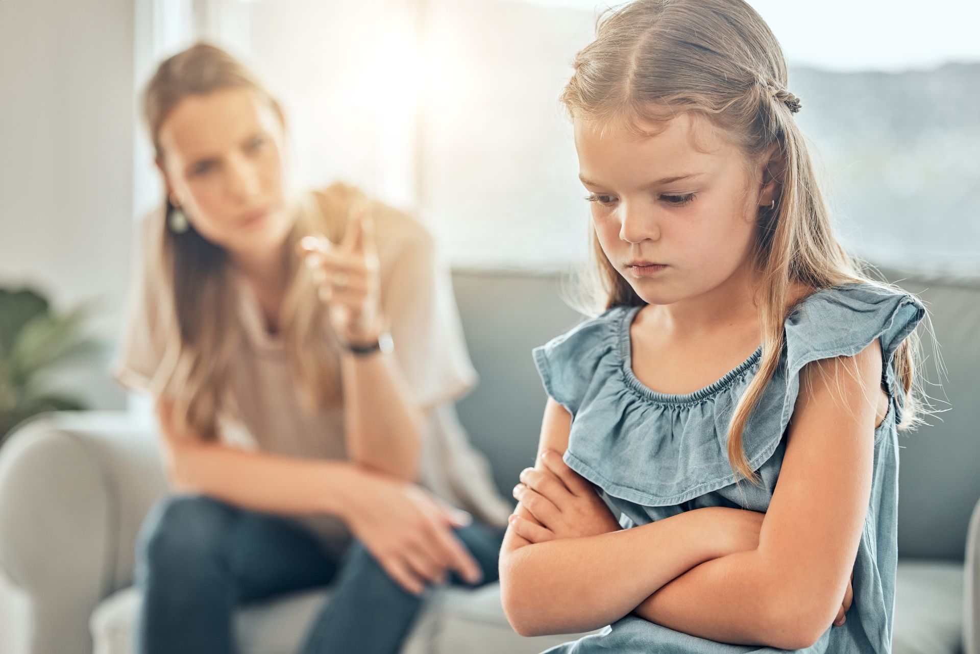 Girl with crossed arms, looking upset as woman points, blurred in background.