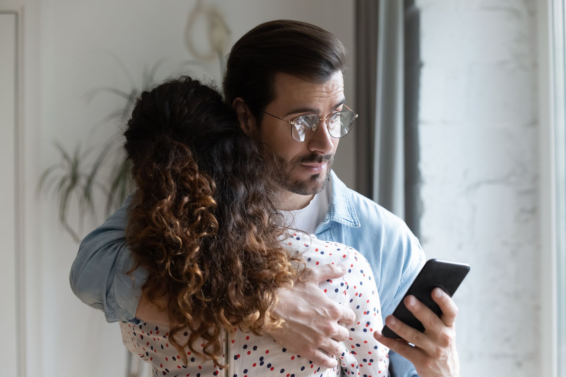 Man hugging woman while looking at a phone in front of a window.