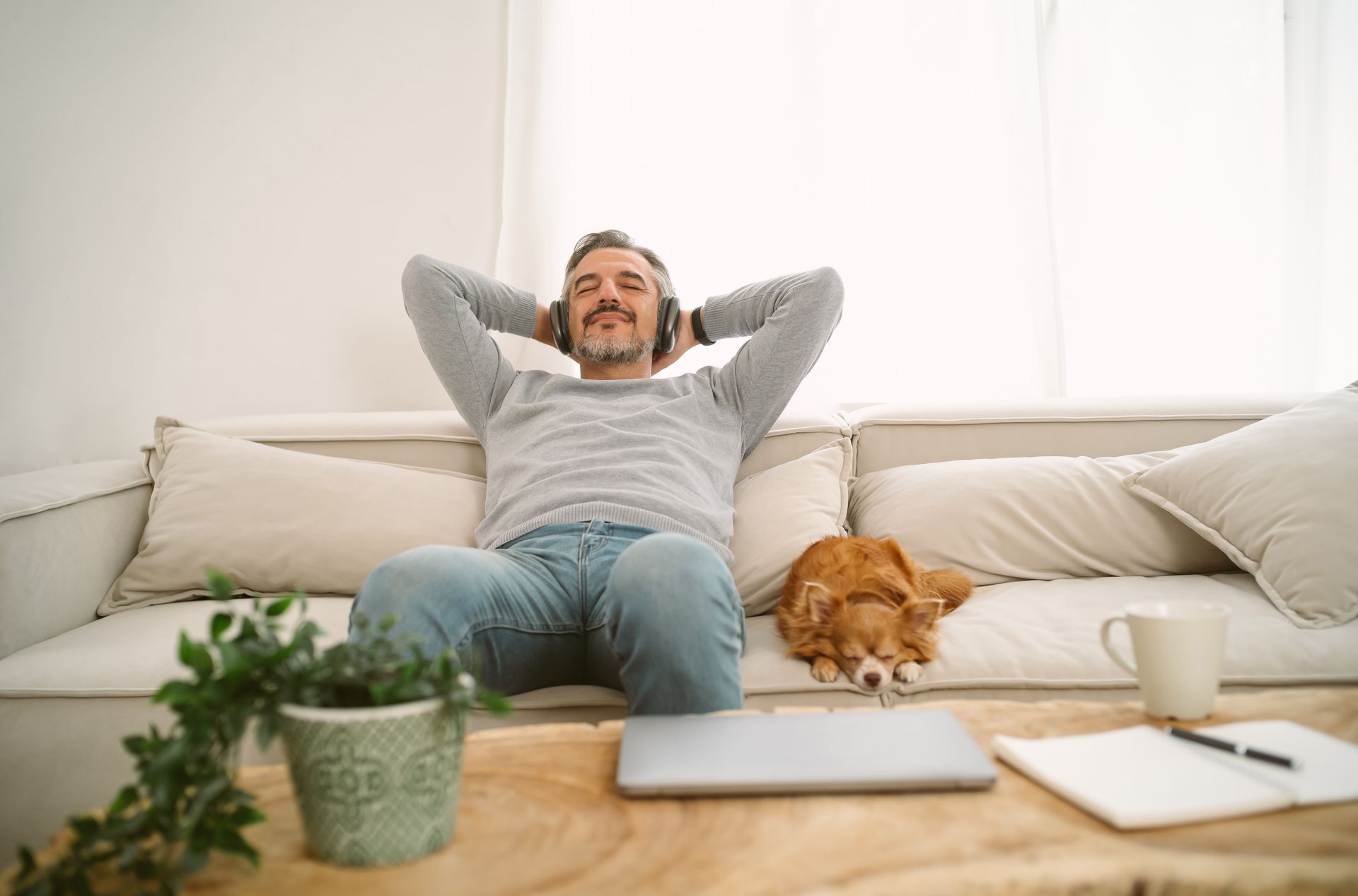 Man relaxing on a sofa, wearing headphones, with dog; laptop and notepad on the table.
