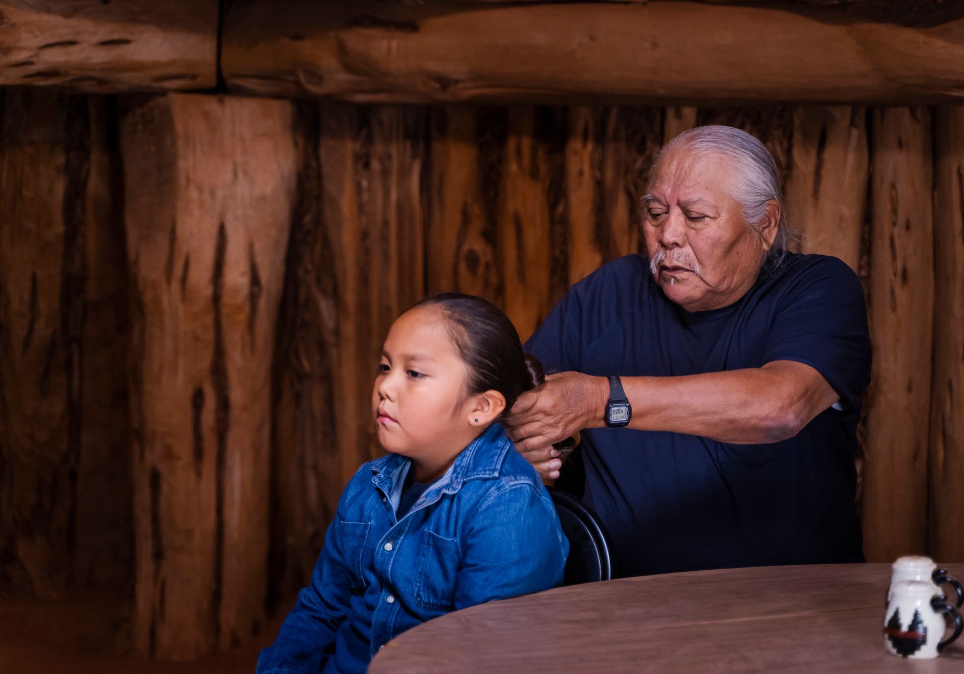 Man braiding girl's hair indoors, possibly a dwelling. Both are at a table, wooden walls visible.