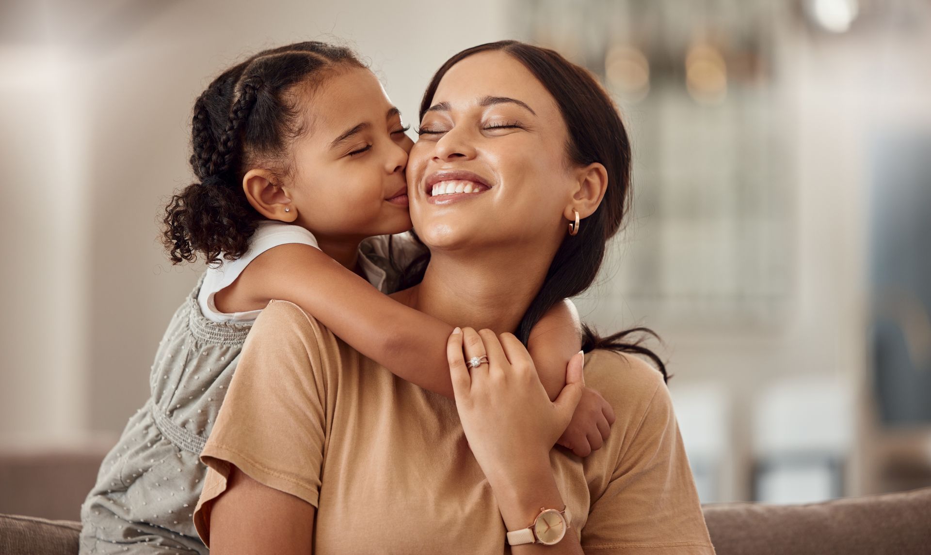 A young child kisses a smiling woman's cheek; both are embracing indoors.