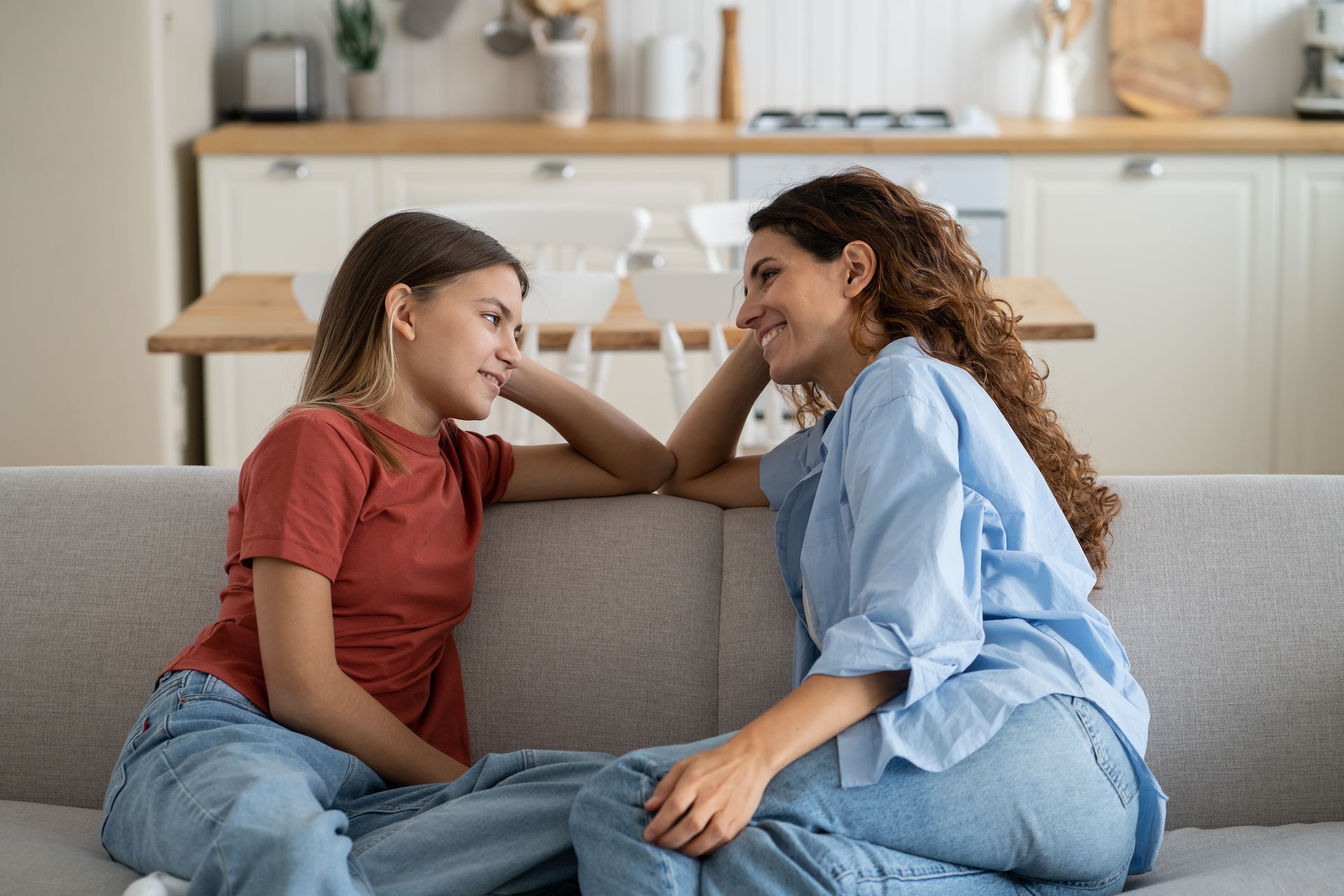 Woman and teen girl sit close on a sofa, looking at each other, in a light-filled kitchen.