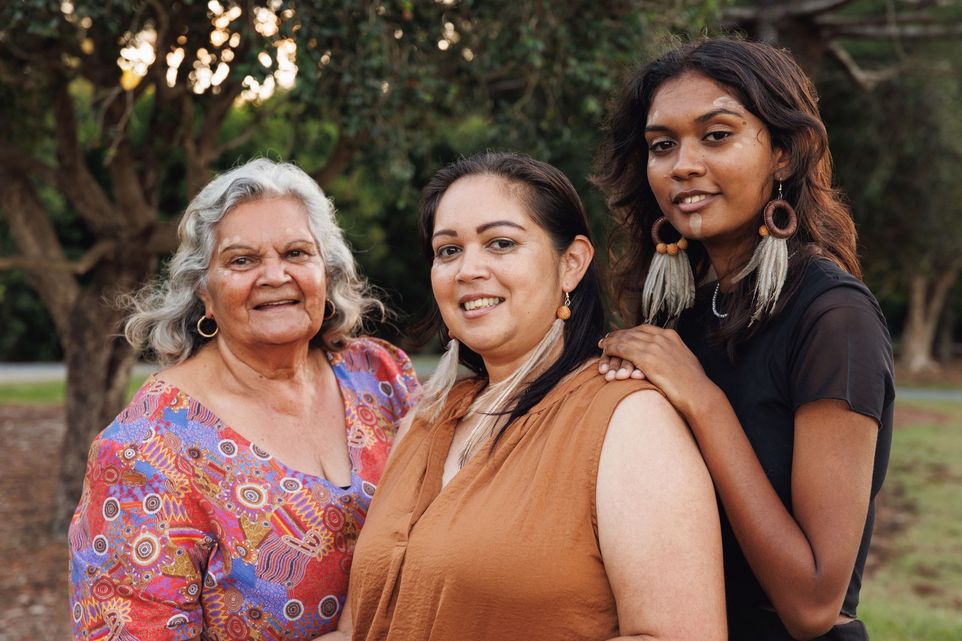 Three women in park; older woman in patterned top, younger in brown, youngest in black top; smiling.