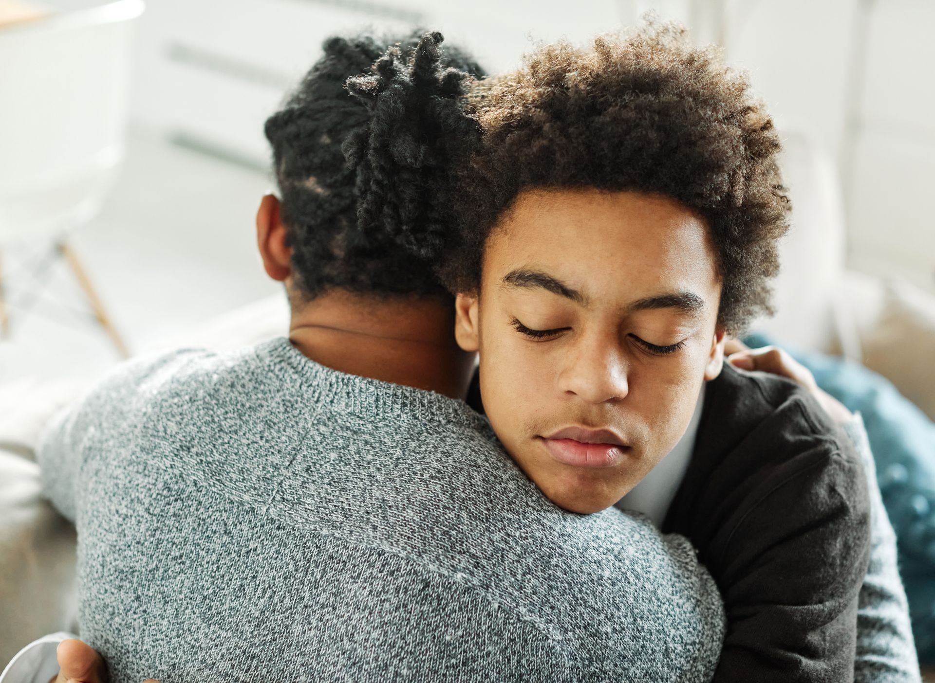 Two people embracing indoors; one with closed eyes, resting head on the other's shoulder.