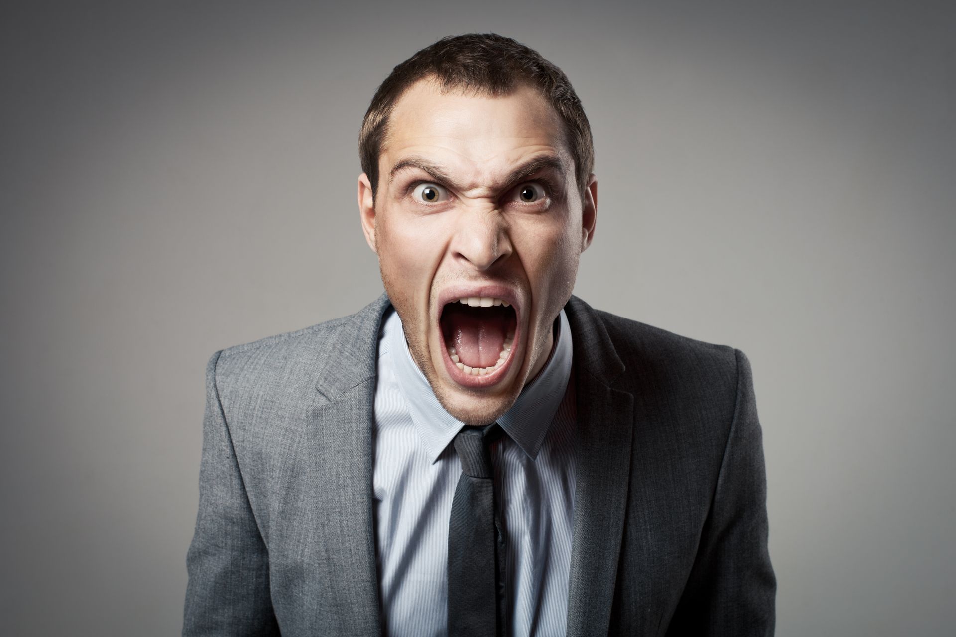 Man in a suit, screaming with mouth wide open; angry expression on a gray background.