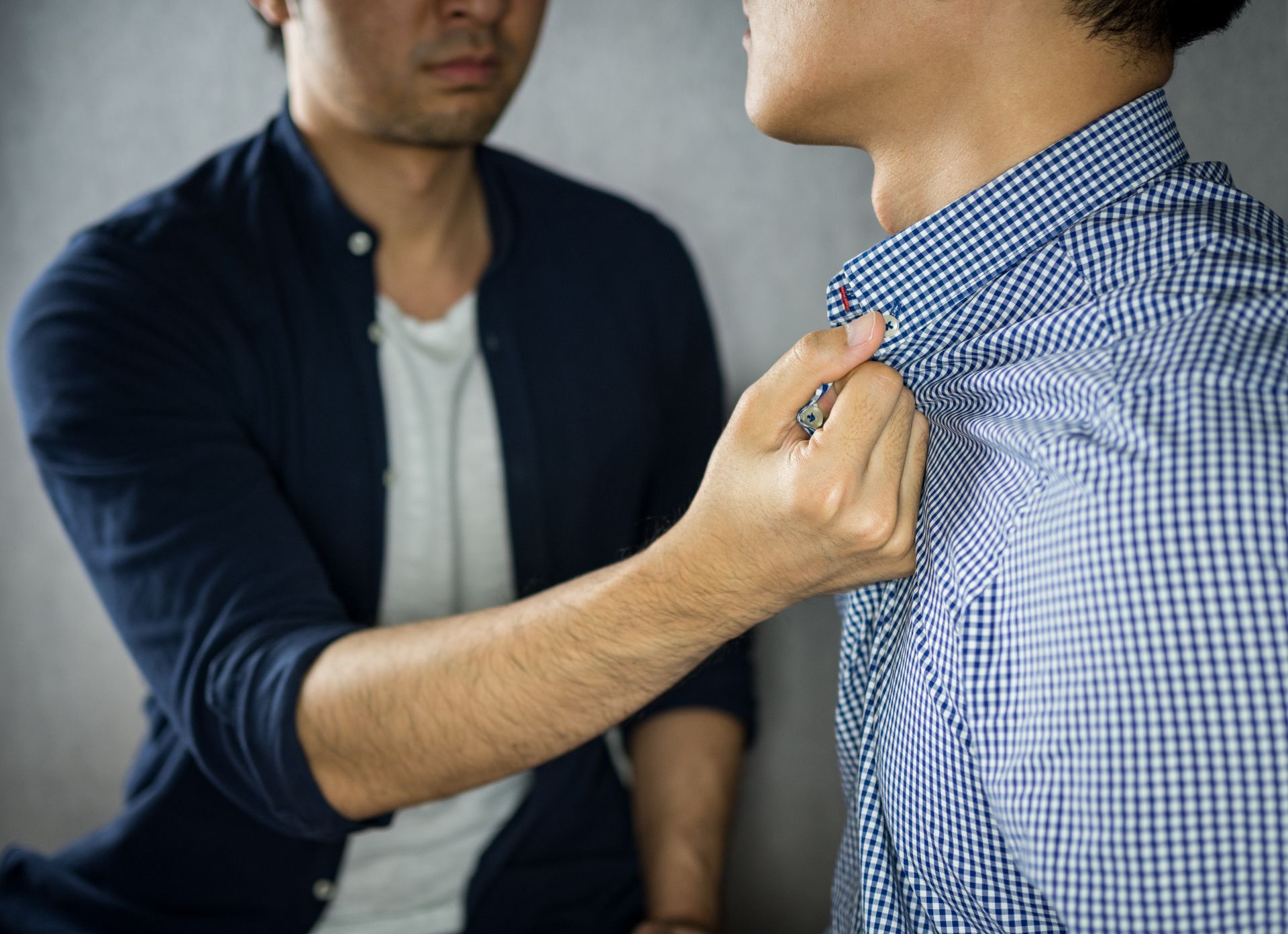 Man's hand gripping another man's shirt collar, face showing anger, near a gray wall.