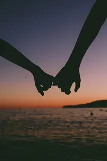 Silhouette of two hands, pinky fingers touching against a sunset over the water.