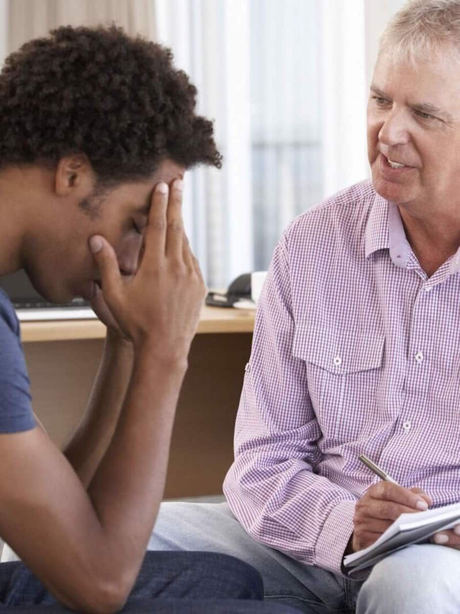 Person with head in hands, sitting across from a person taking notes, indoors.