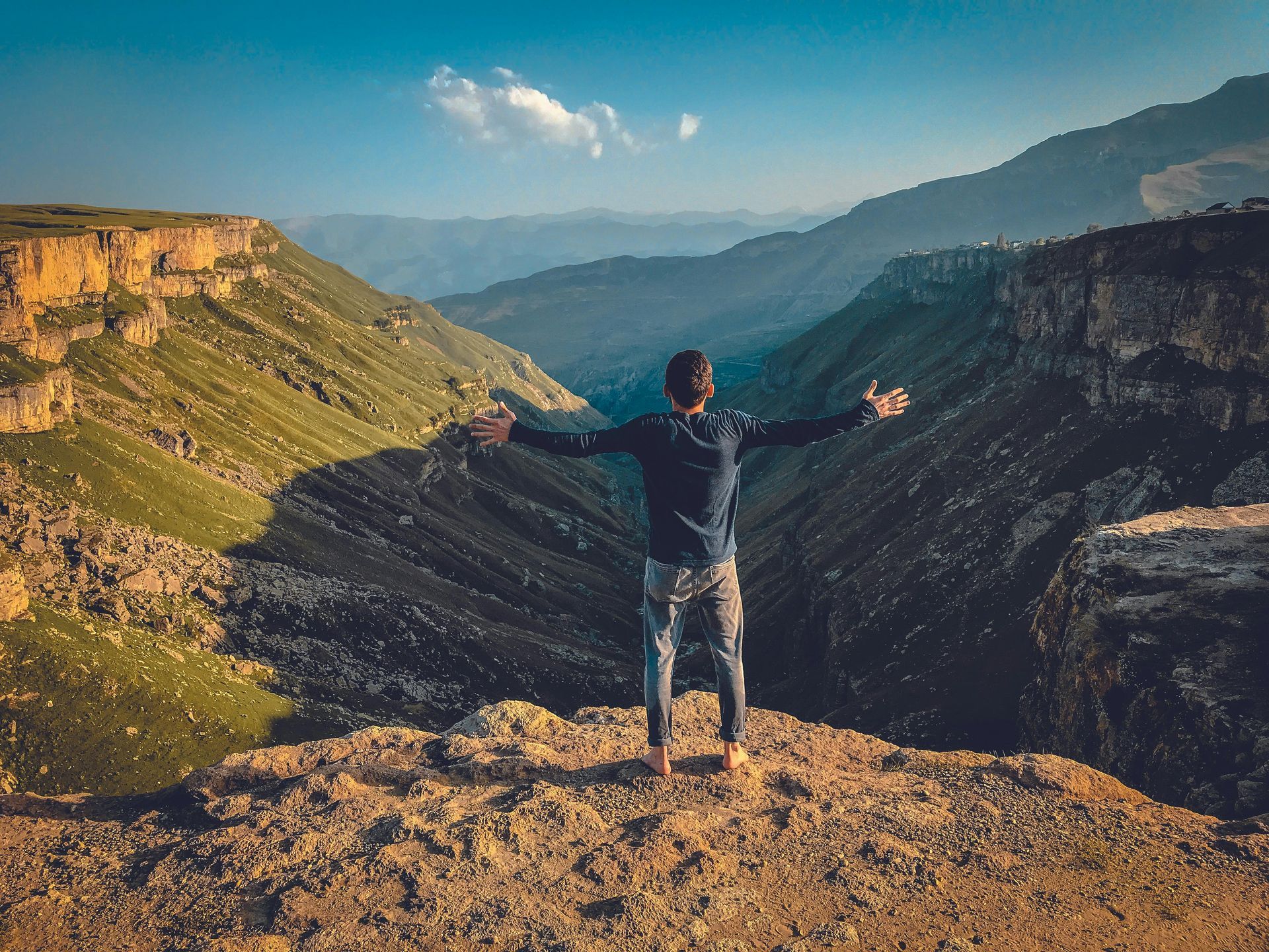 Man with arms outstretched on cliff overlooking a valley. Sunlight, mountains, blue sky.