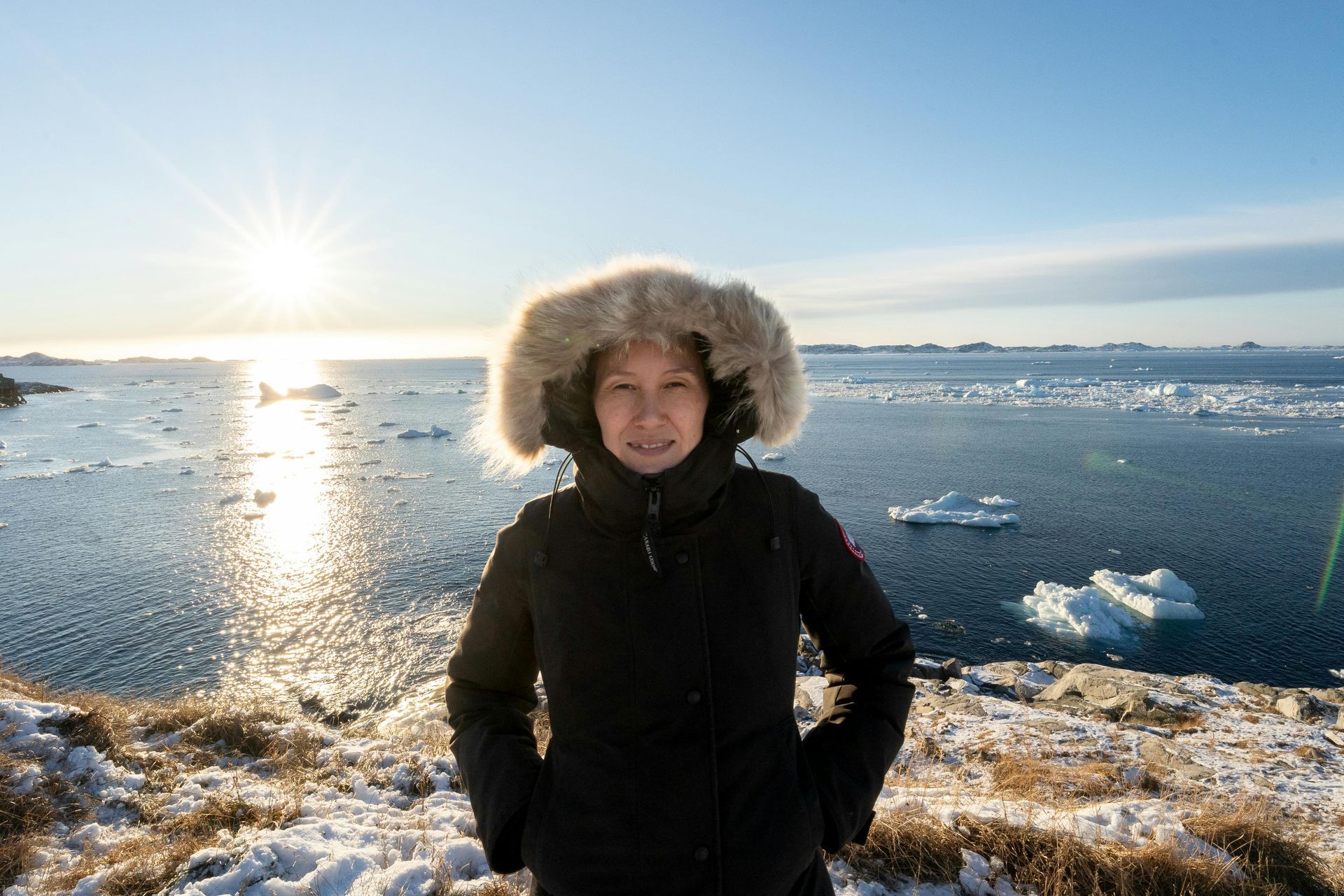 Person in a fur-lined hooded coat standing by a snowy, icy shoreline at sunrise