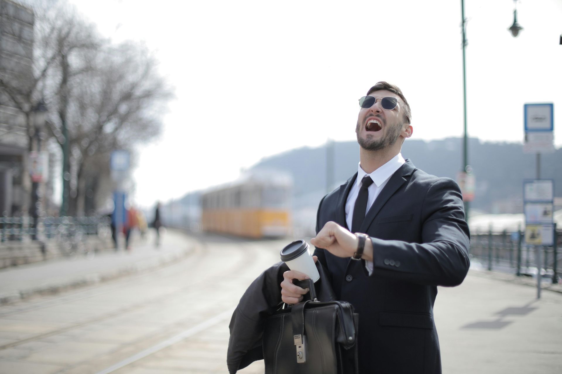 Man in suit, sunglasses checks watch, holds briefcase and coffee on city street with tram.