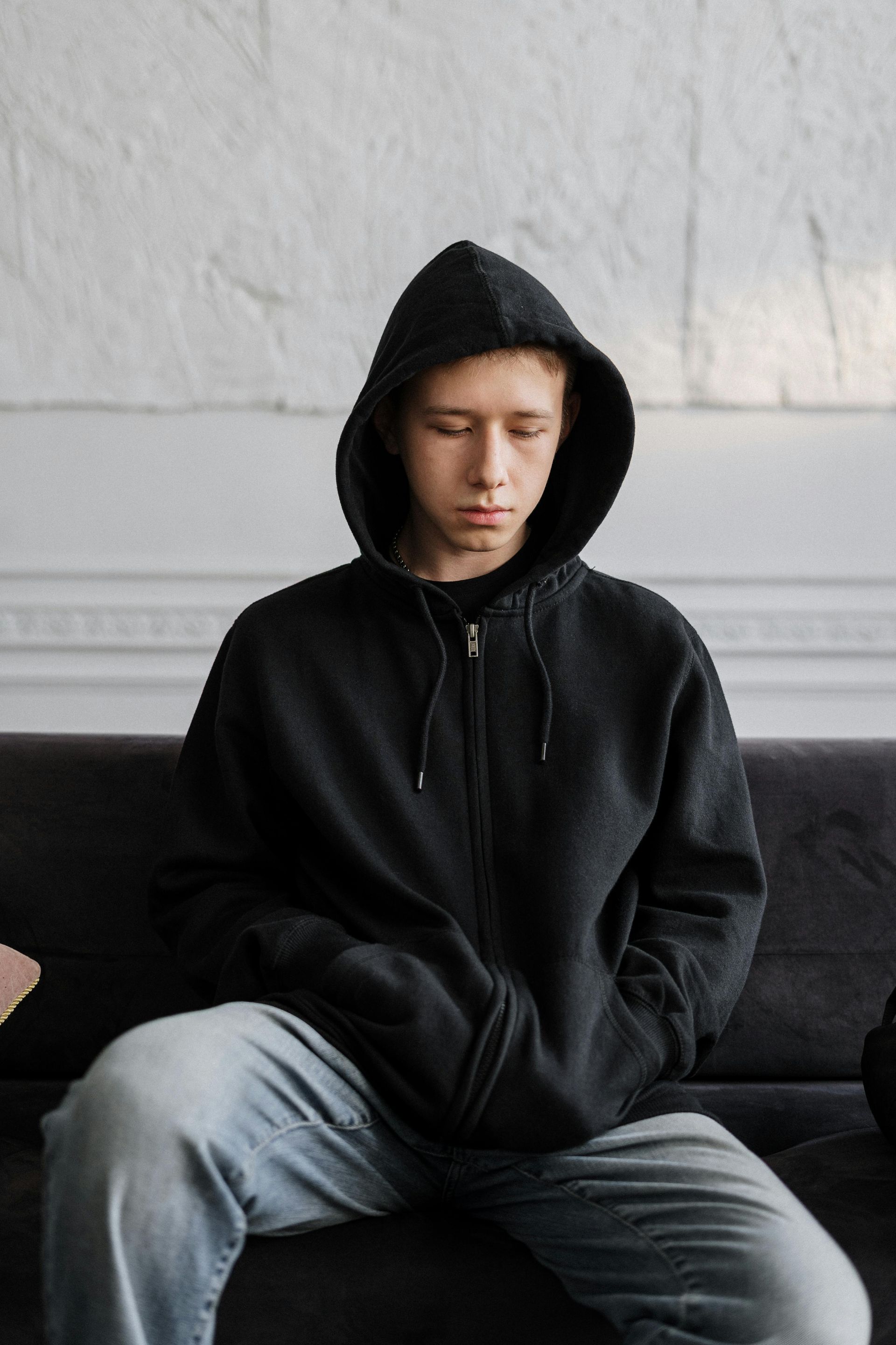 Person in black hoodie with hood up, sitting on a couch, looking down.