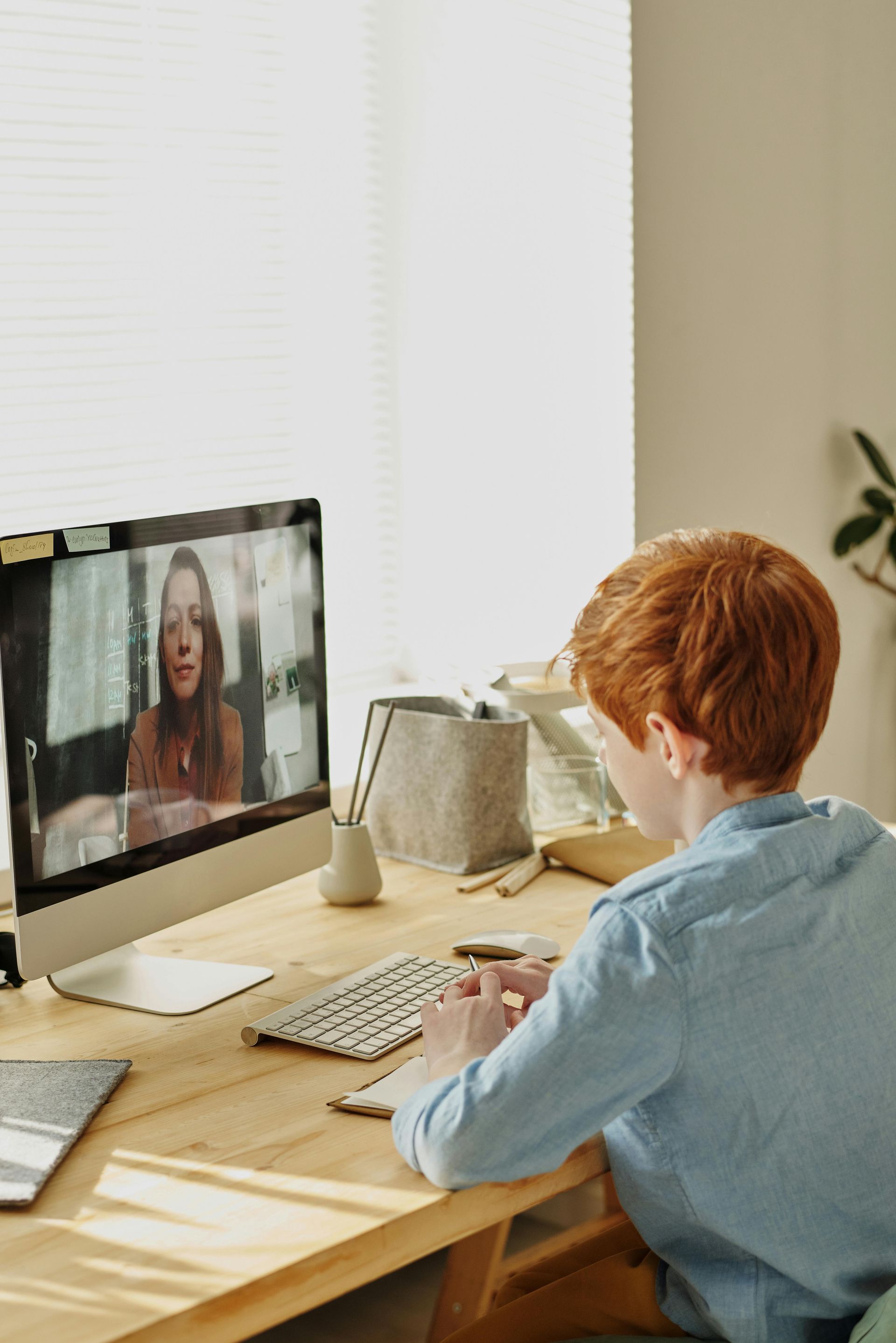 A person with red hair sits at a desk, looking at a computer screen showing a video call with a woman.