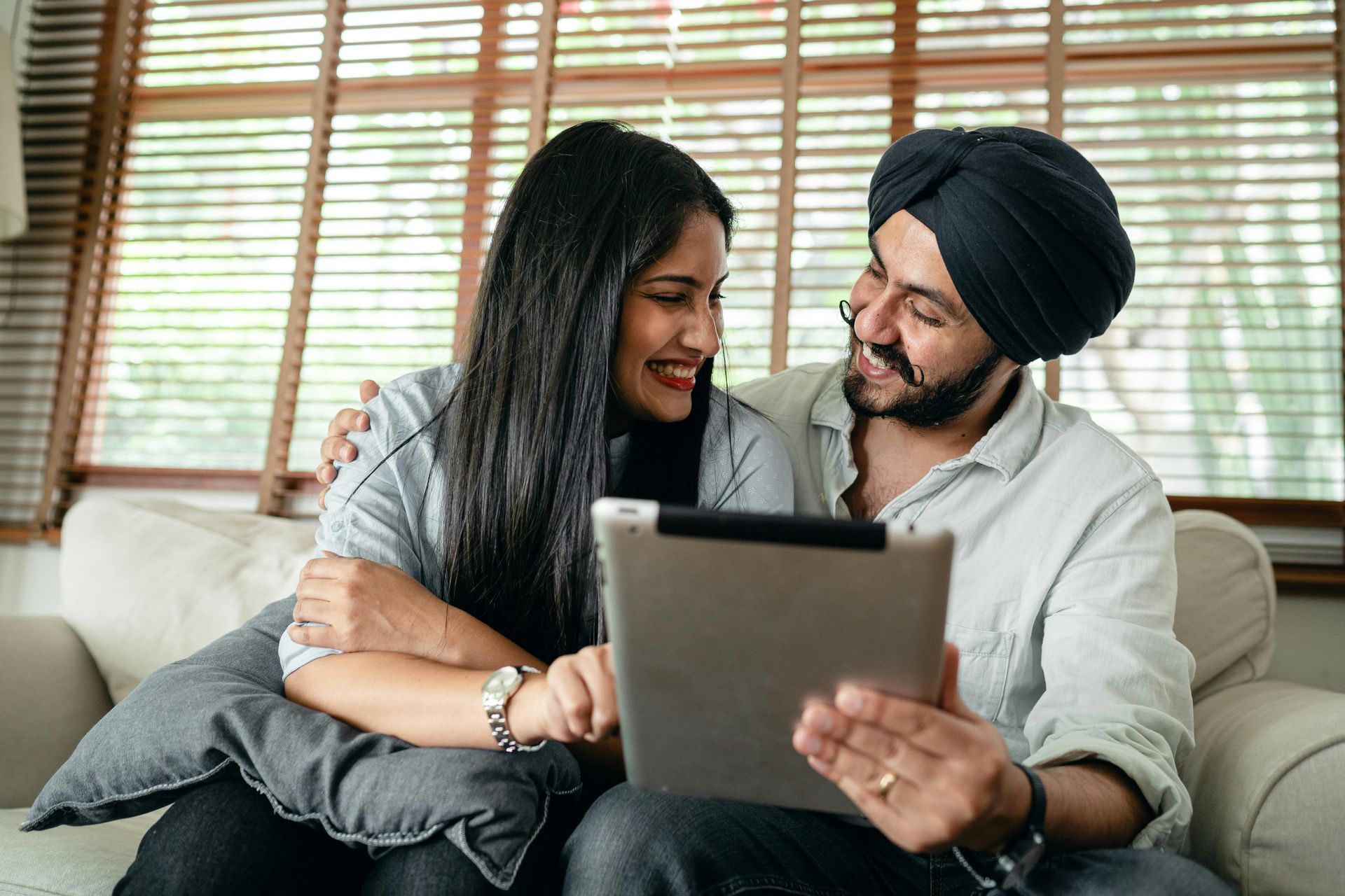 Couple on a couch, looking at a tablet, smiling. They are indoors.
