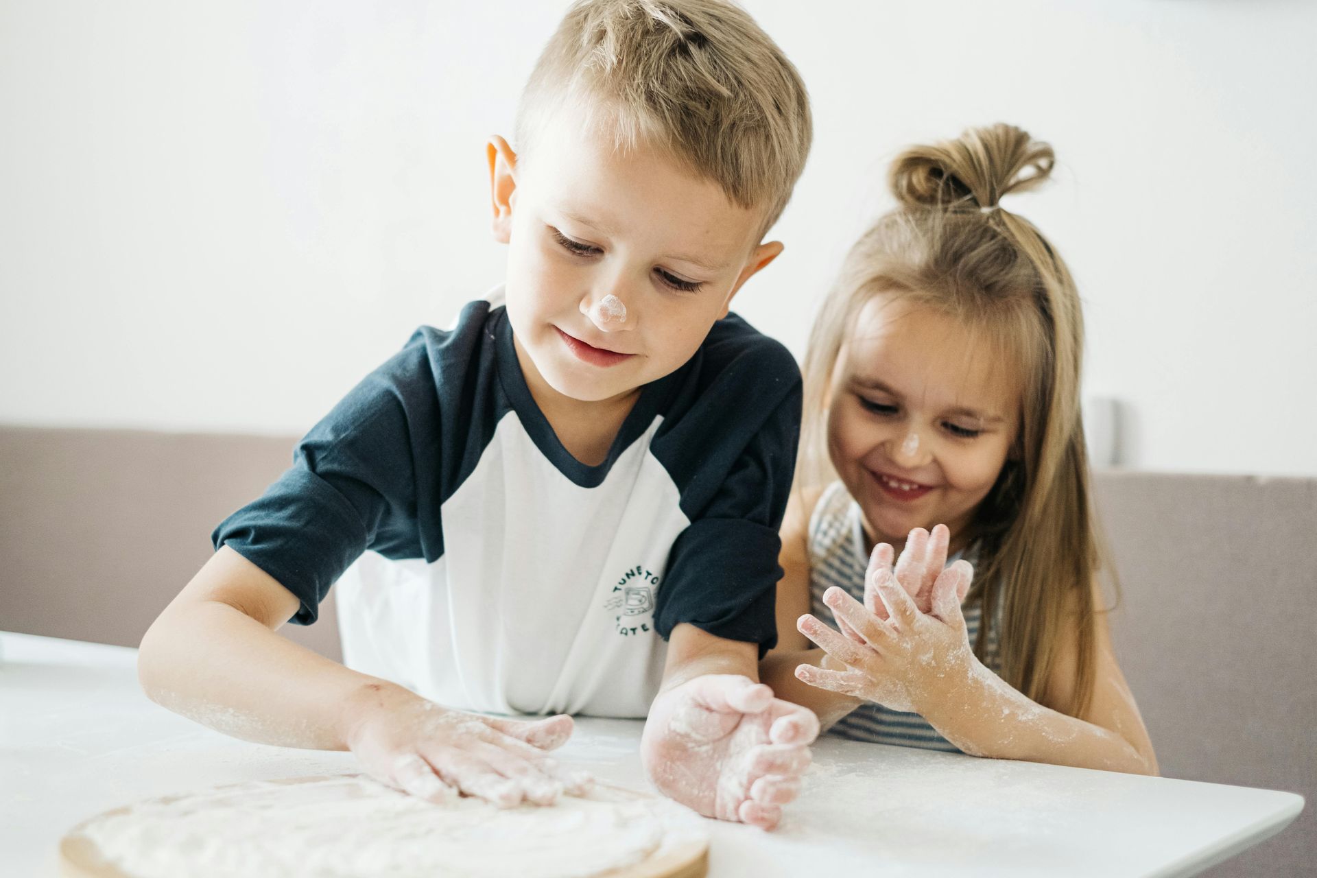 Two children smile as they play with flour on a table. One rolls the flour while the other claps hands.