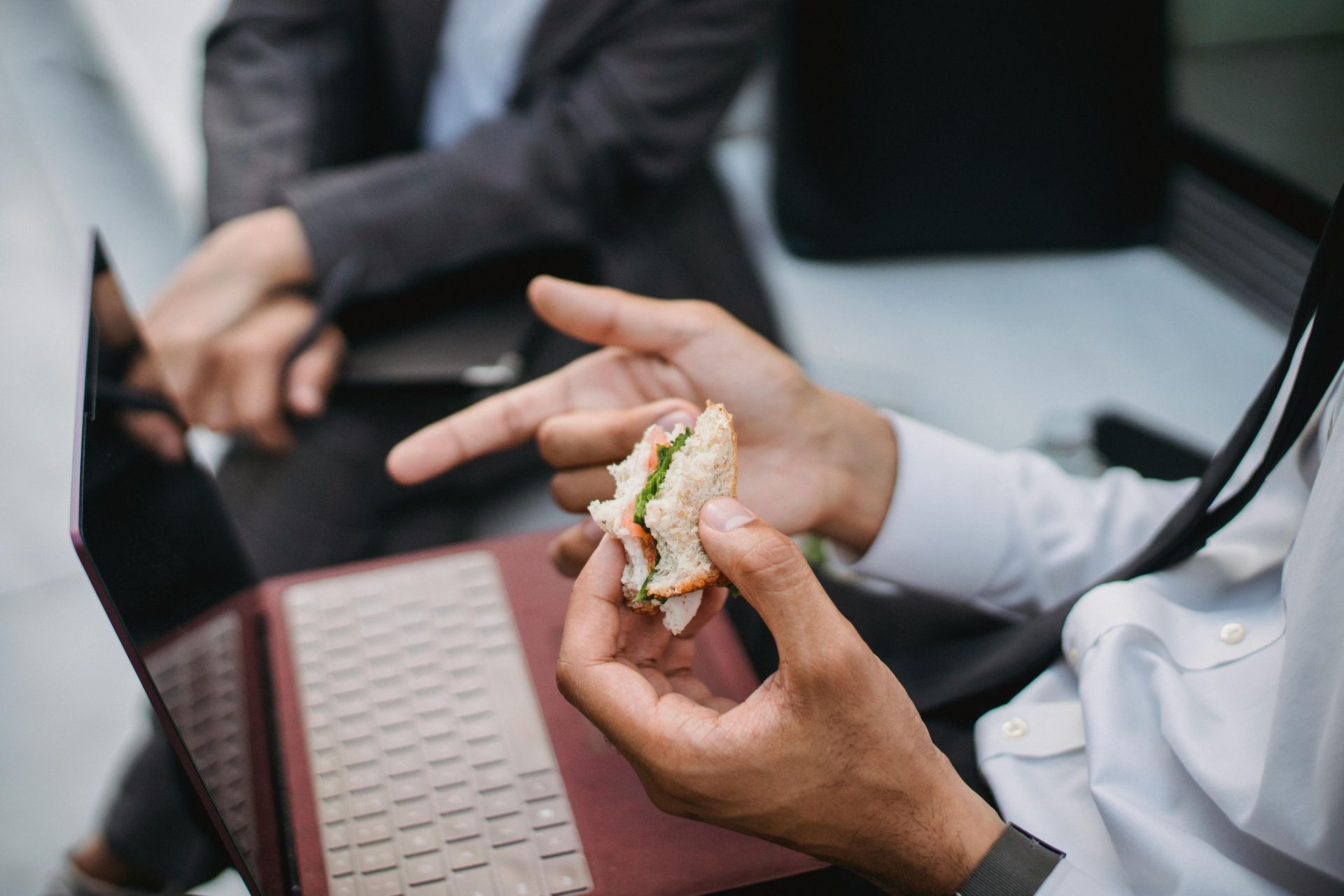 Person in suit eats a sandwich, pointing at laptop while sitting near another person.