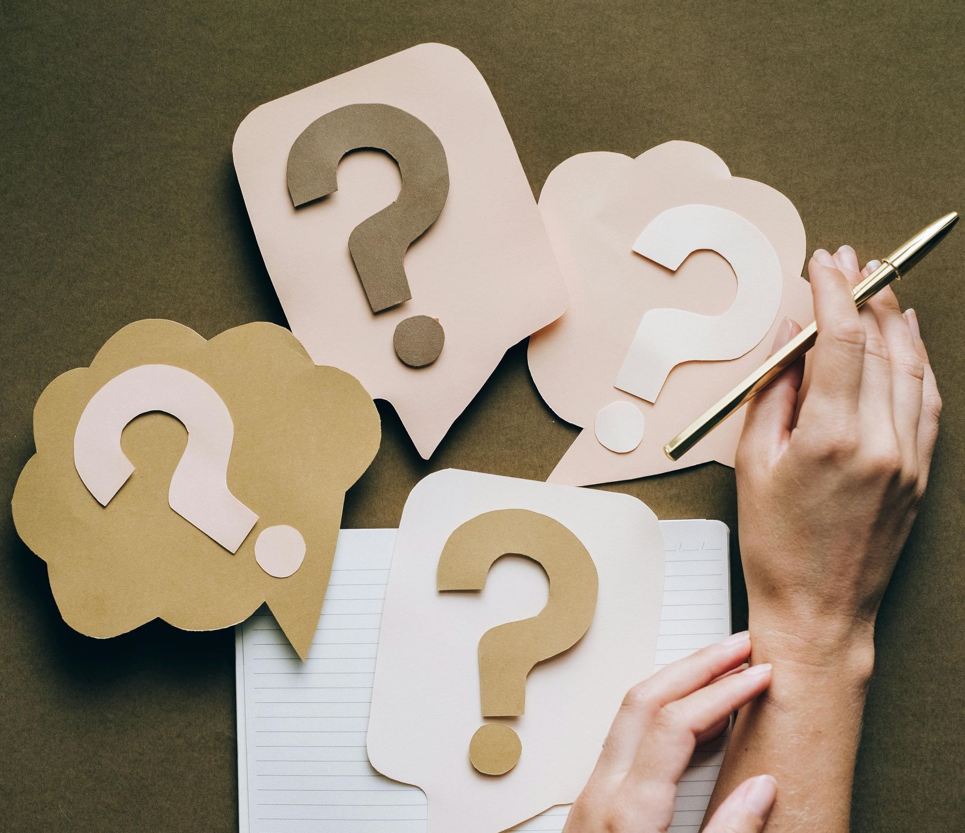 Hands with a pen, surrounded by paper question mark cutouts, on a brown surface.