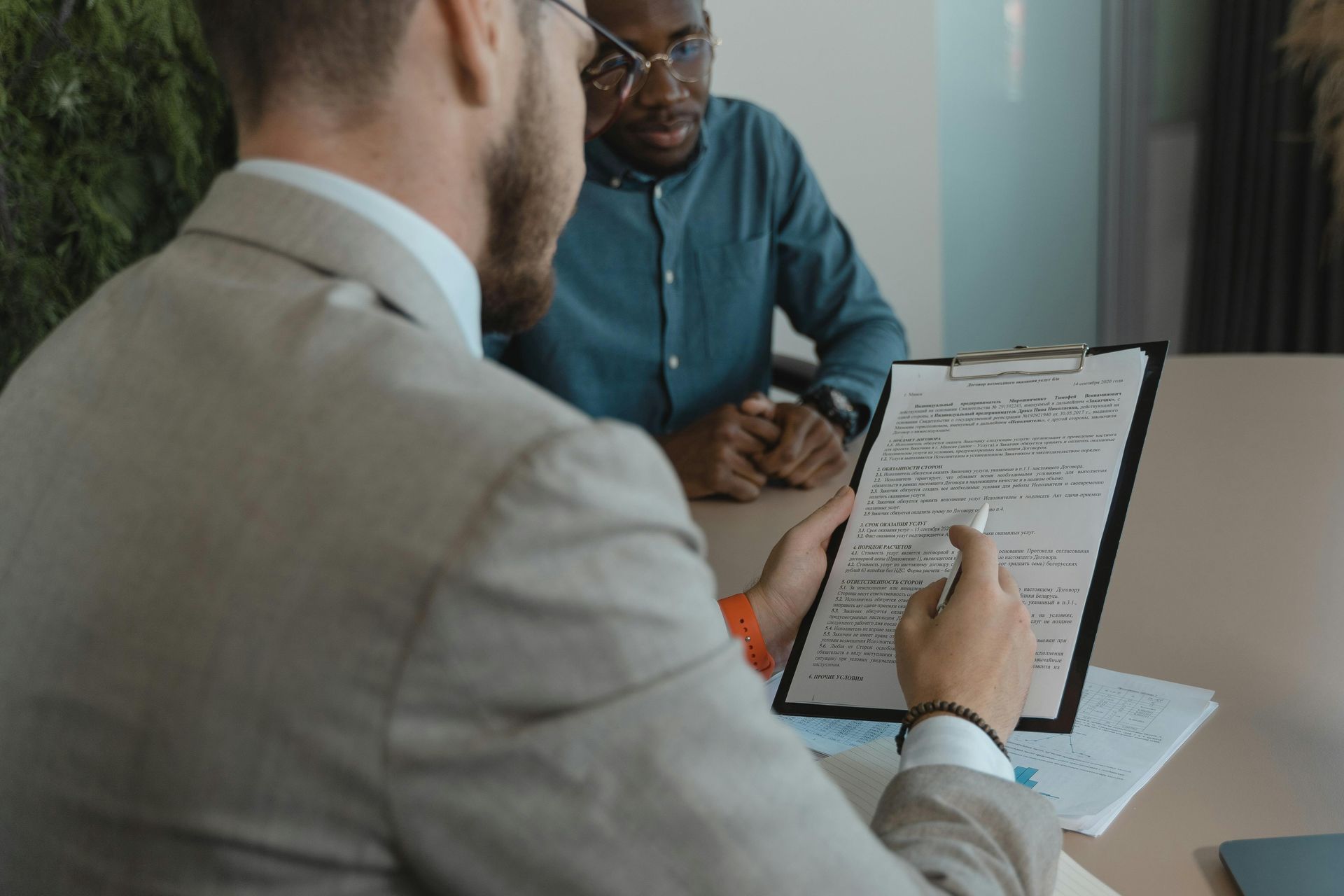 Man in tan suit pointing at document on clipboard, sitting across from another man in blue shirt.