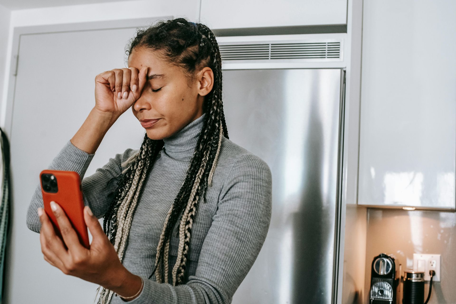 Woman holding orange phone, hand on forehead, looking down, in kitchen with stainless steel fridge.