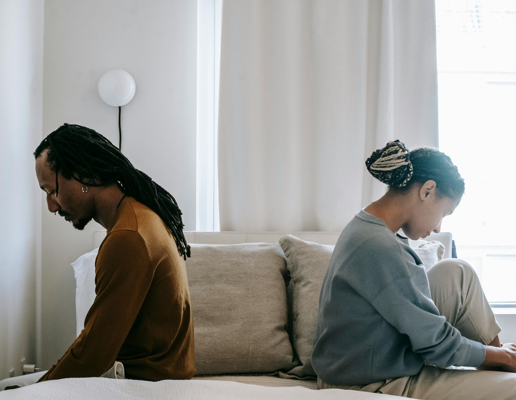 Couple sitting on a bed, backs turned to each other, appearing sad in a bedroom.