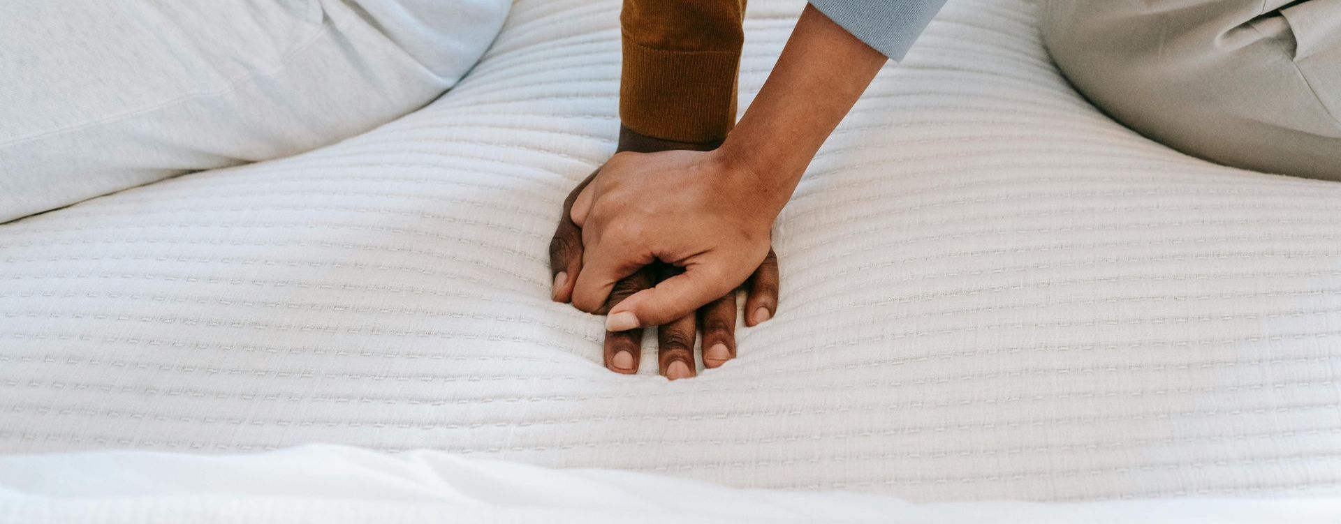 Two hands, one dark-skinned and one light-skinned, clasped together on a white mattress.