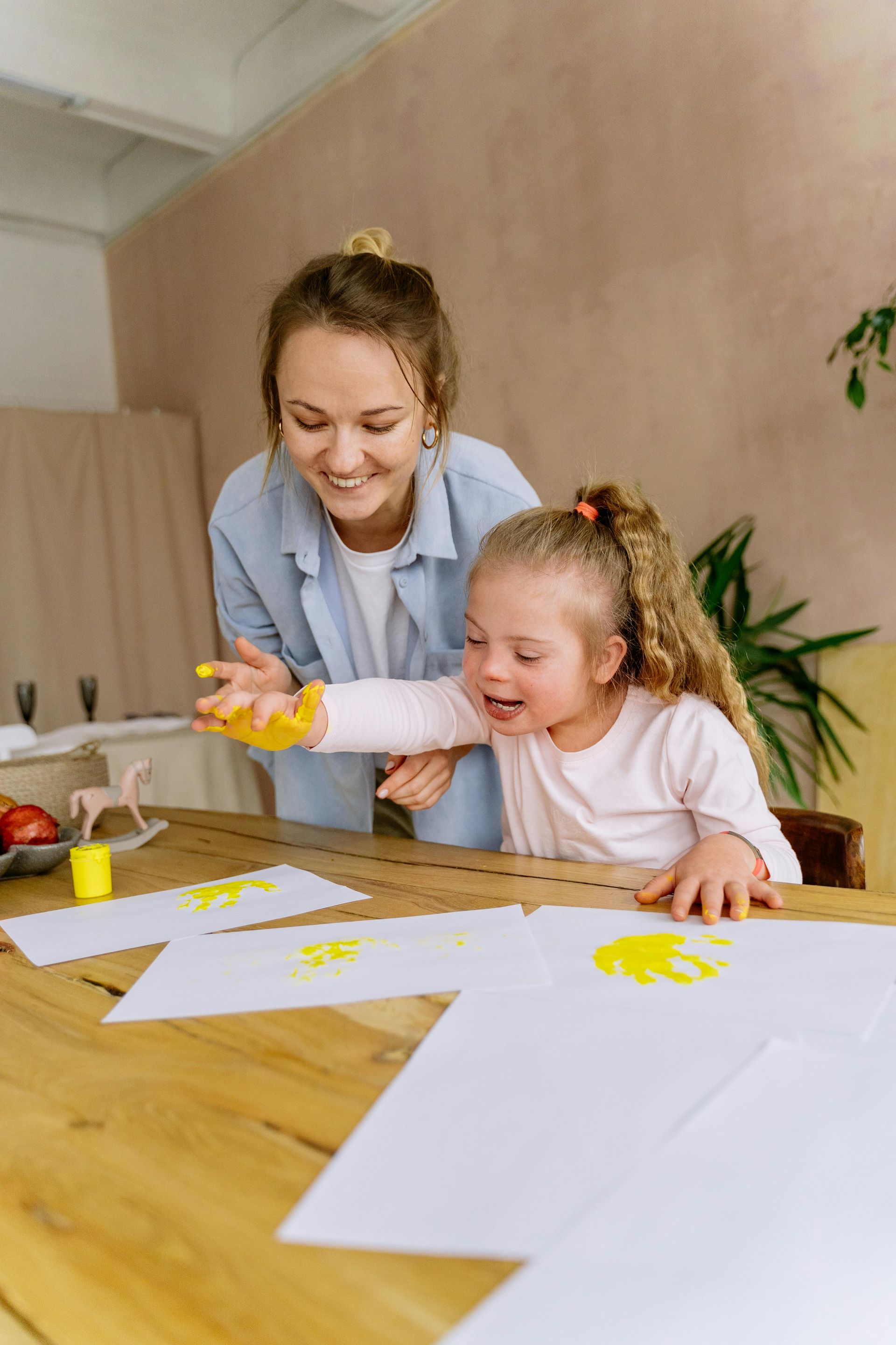 Woman assisting a child with Down syndrome in painting with yellow paint on paper at a table.