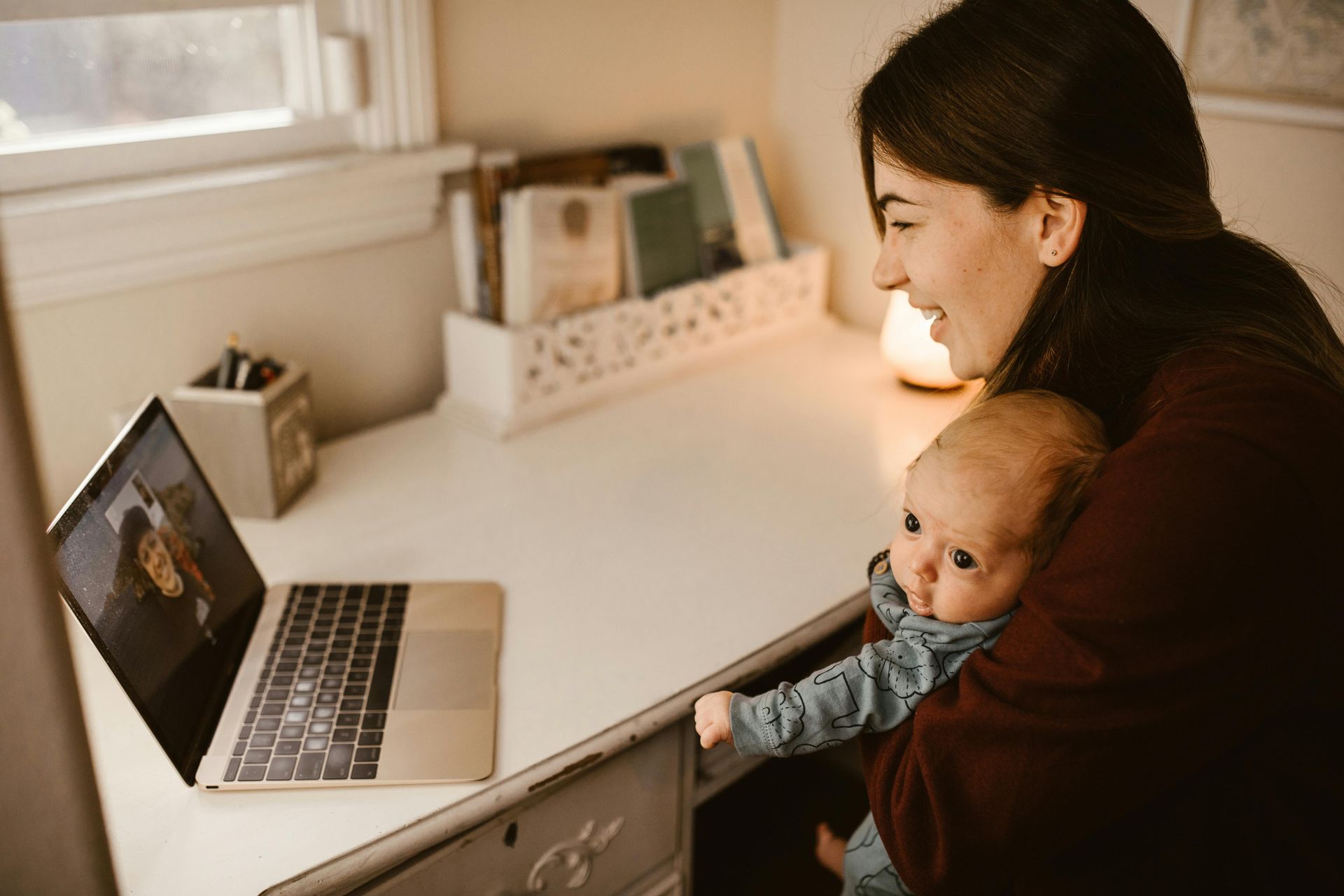 Woman holding a baby smiles at laptop screen on a desk; soft lighting, book shelf in background.