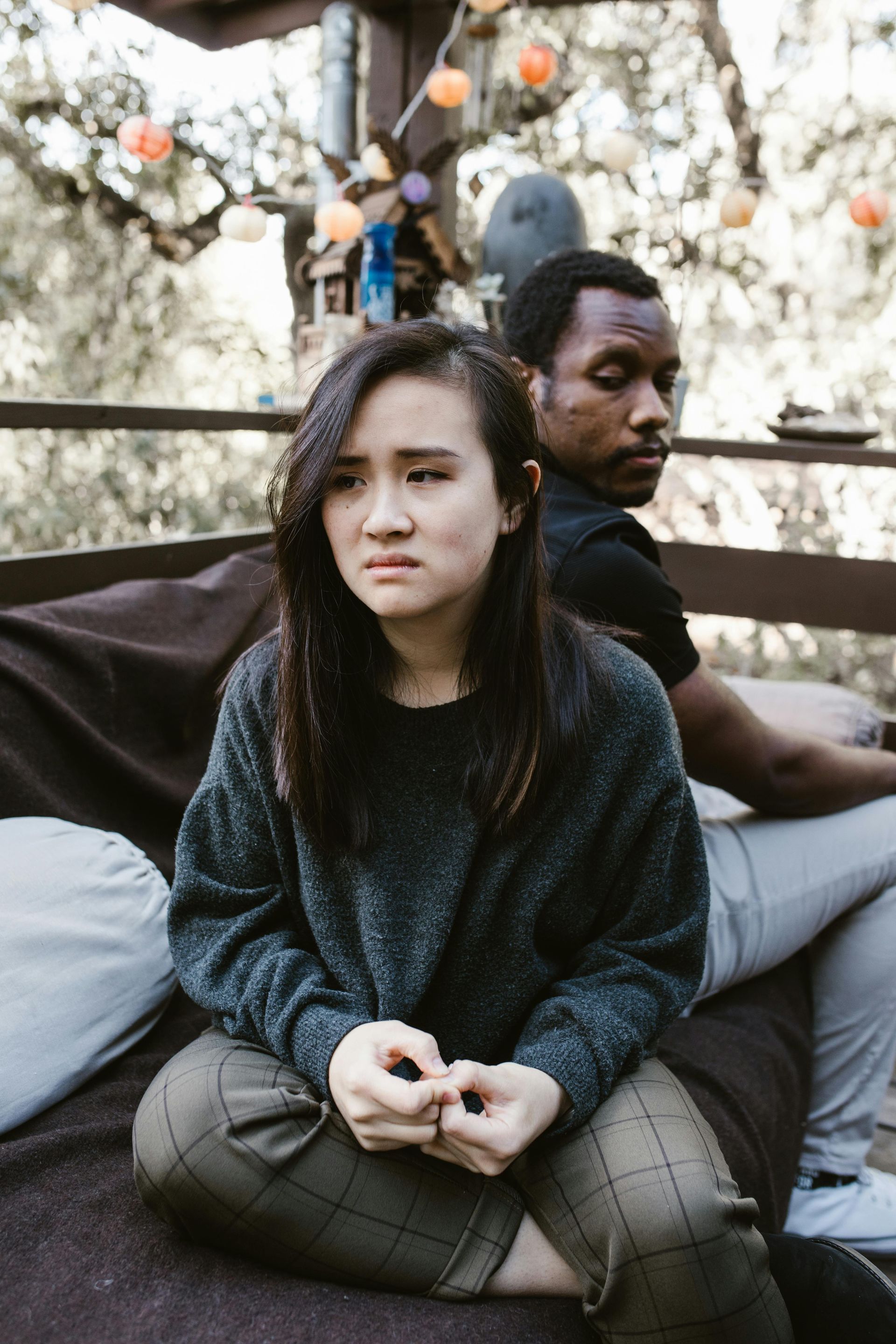 Woman in sweater sits facing forward, looking worried. Man sits behind her, looking away. Outdoors setting.