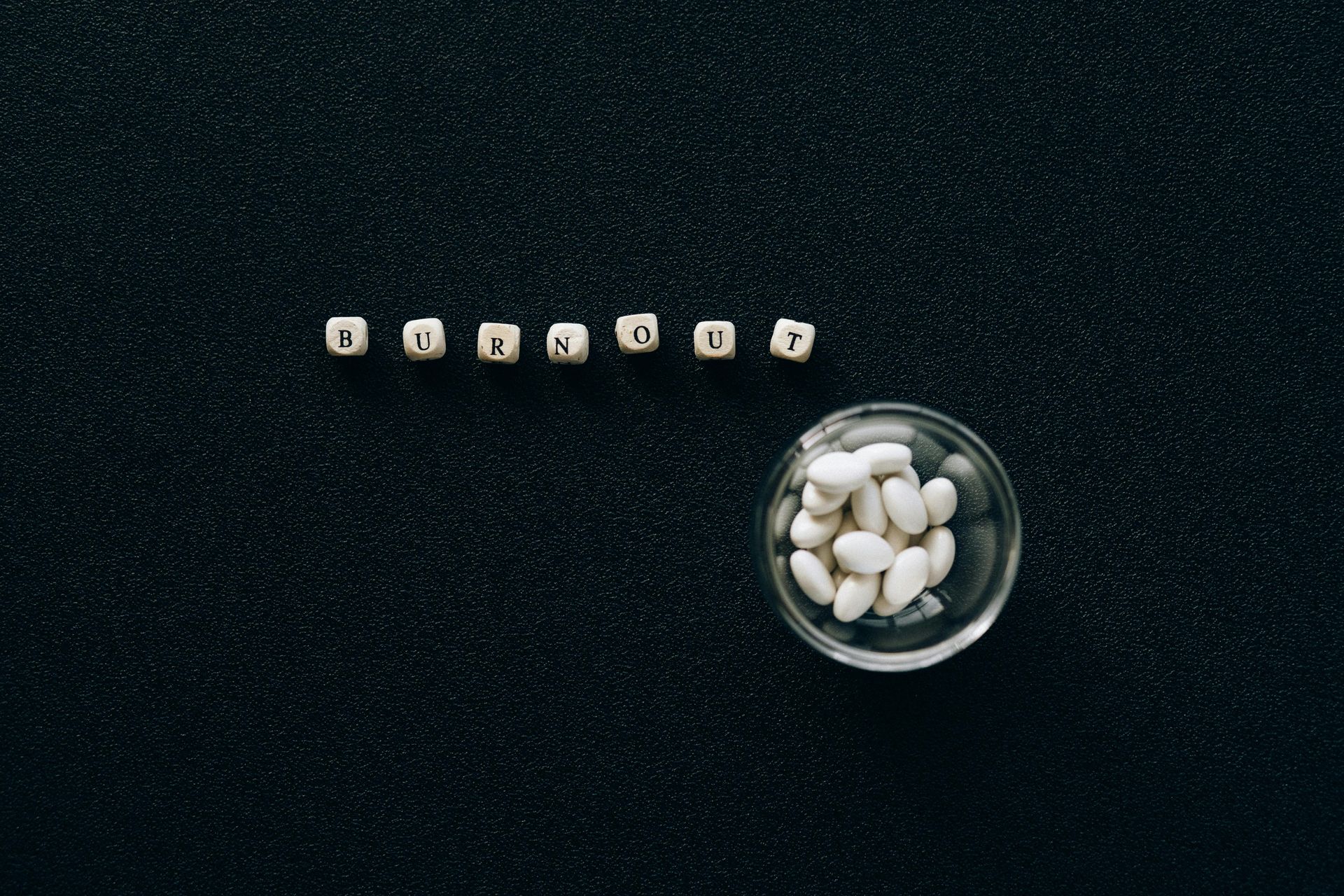 Small wooden cubes are arranged next to a small glass bowl filled with white pills on a black surface.