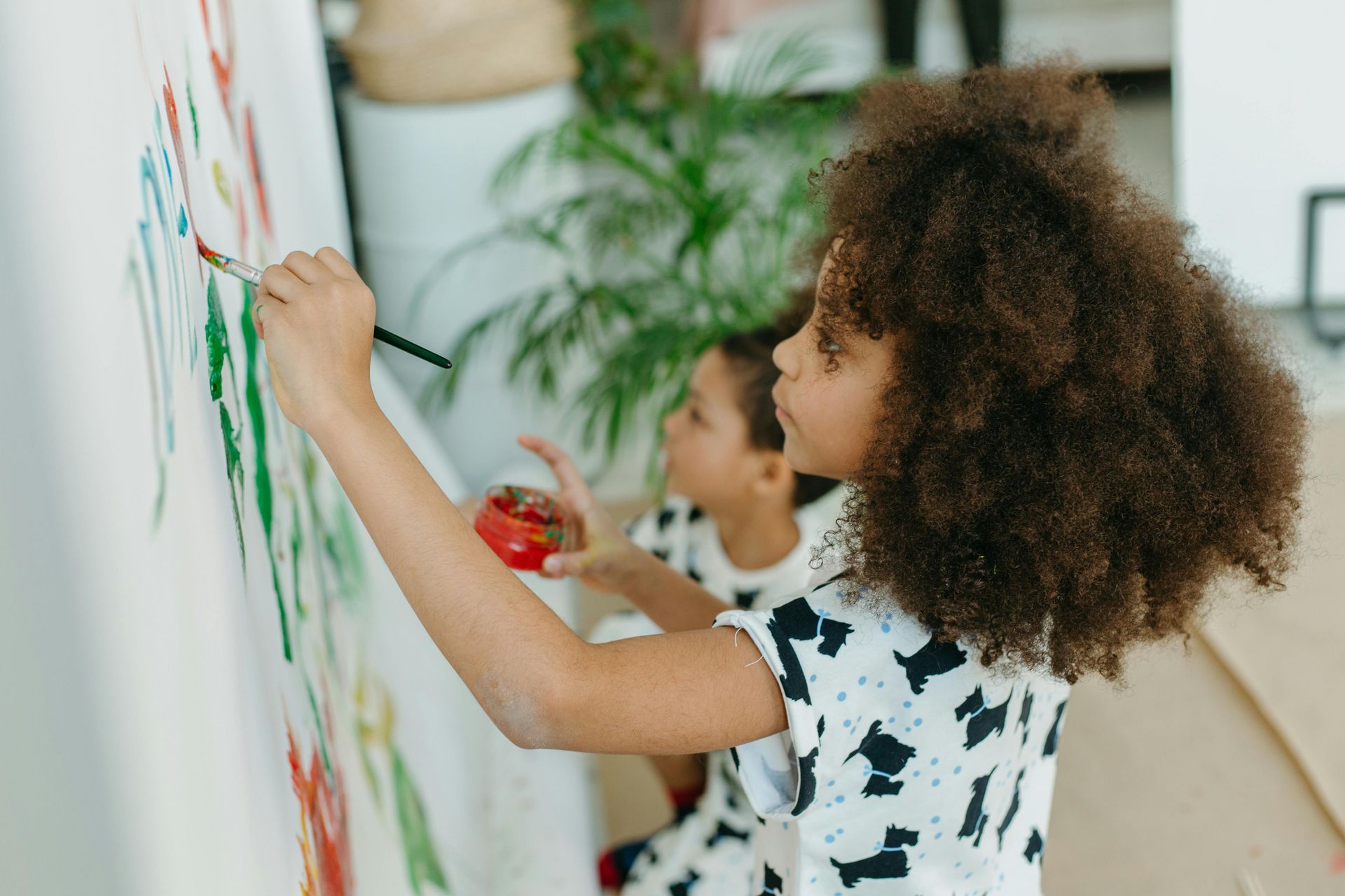 Girl with curly hair painting on a canvas with another child in the background.
