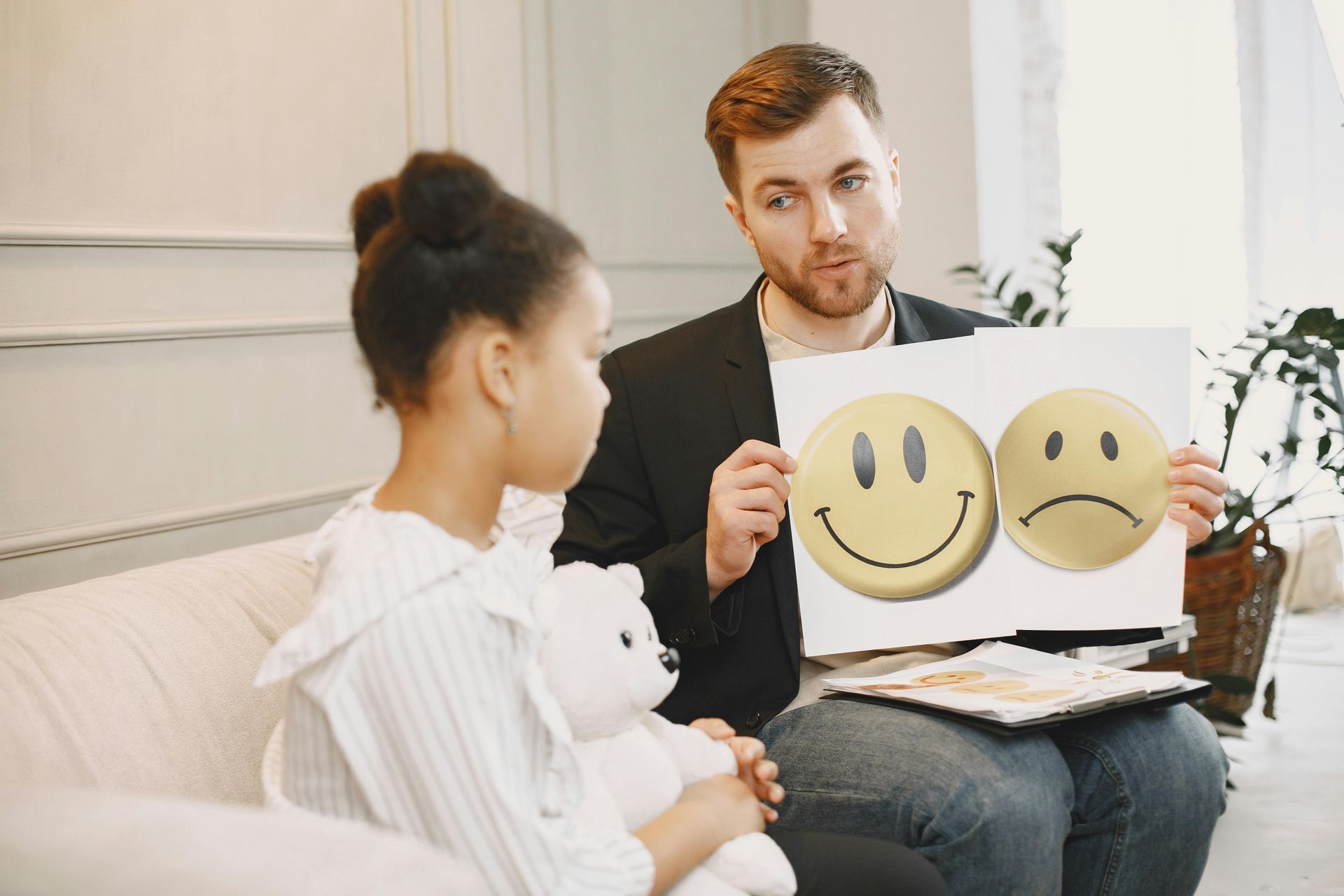 A man holding a drawing of smiley and sad faces, speaking to a child on a couch.