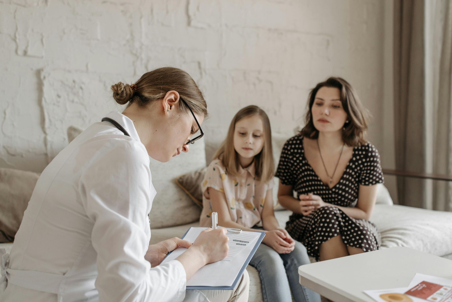 A woman in a white coat writes on a clipboard as a mother and child sit beside her on a couch.