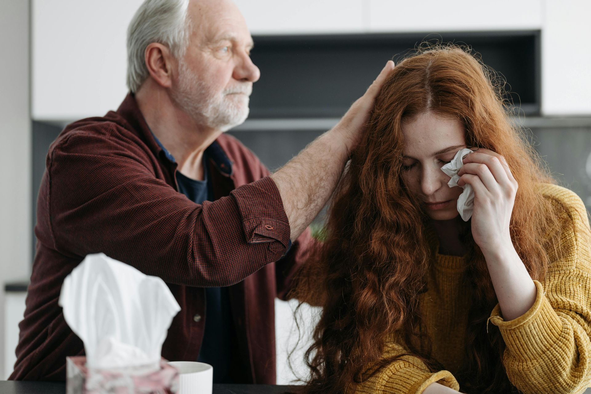 Older person comforts a crying person by placing hand on their head. Tissue box nearby.