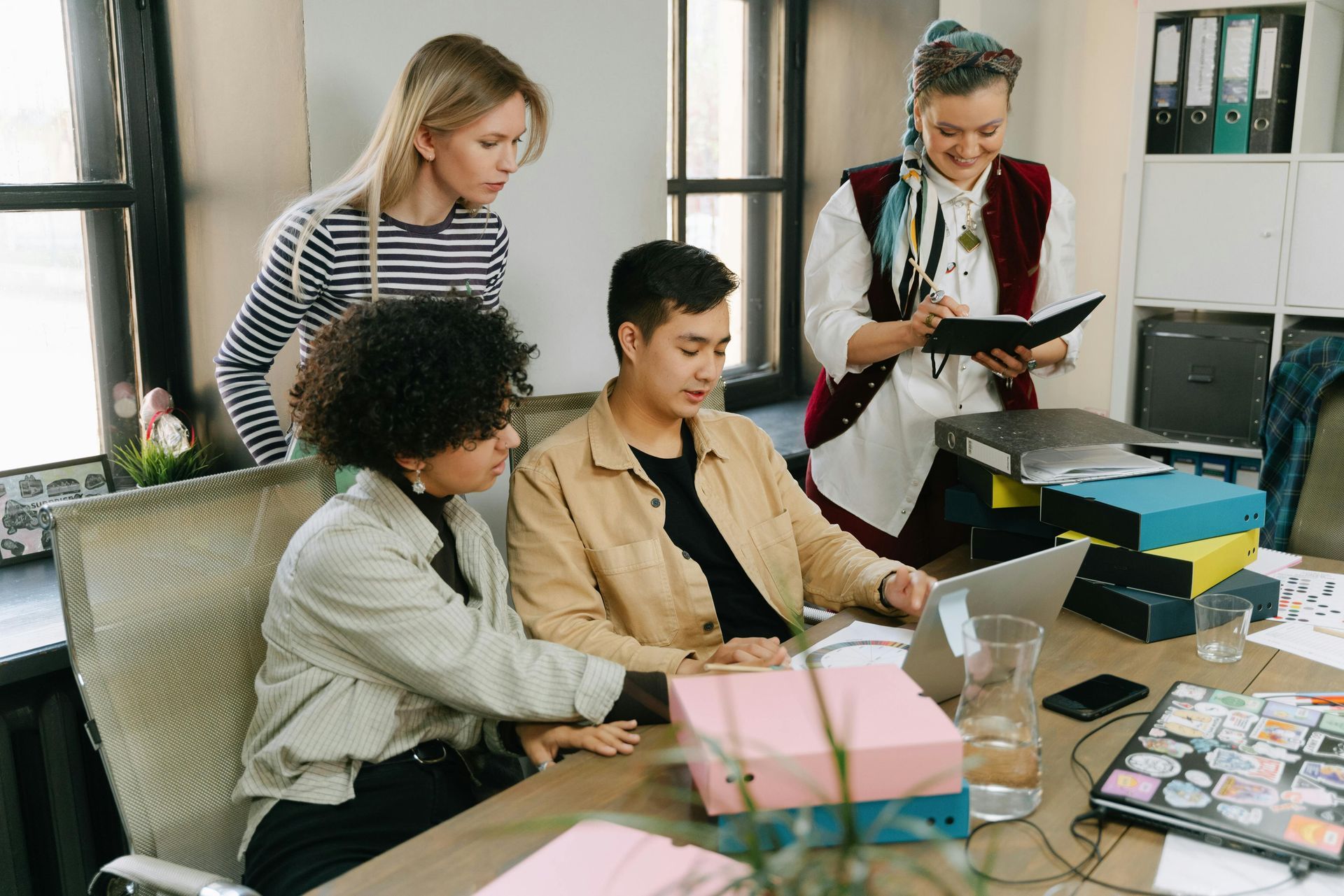 Four people collaborate at an office desk, looking at a laptop and documents. A woman with a clipboard observes.