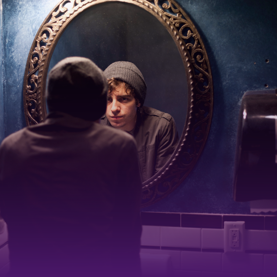 Man looking in a mirror. He wears a beanie and jacket in a dimly lit bathroom.