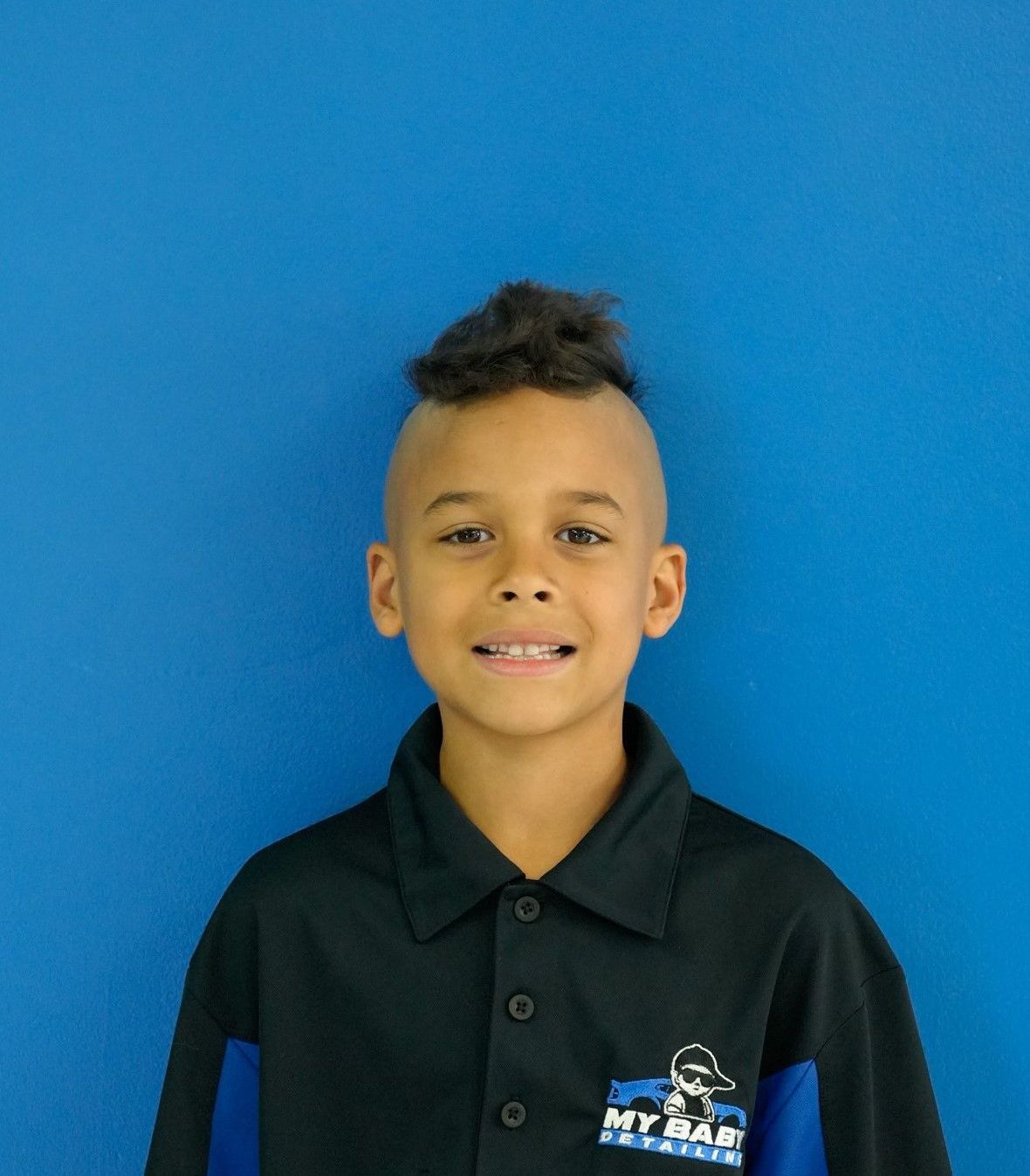 Boy with a dark, textured mohawk and a blue and black collared shirt, smiles in front of a blue background.