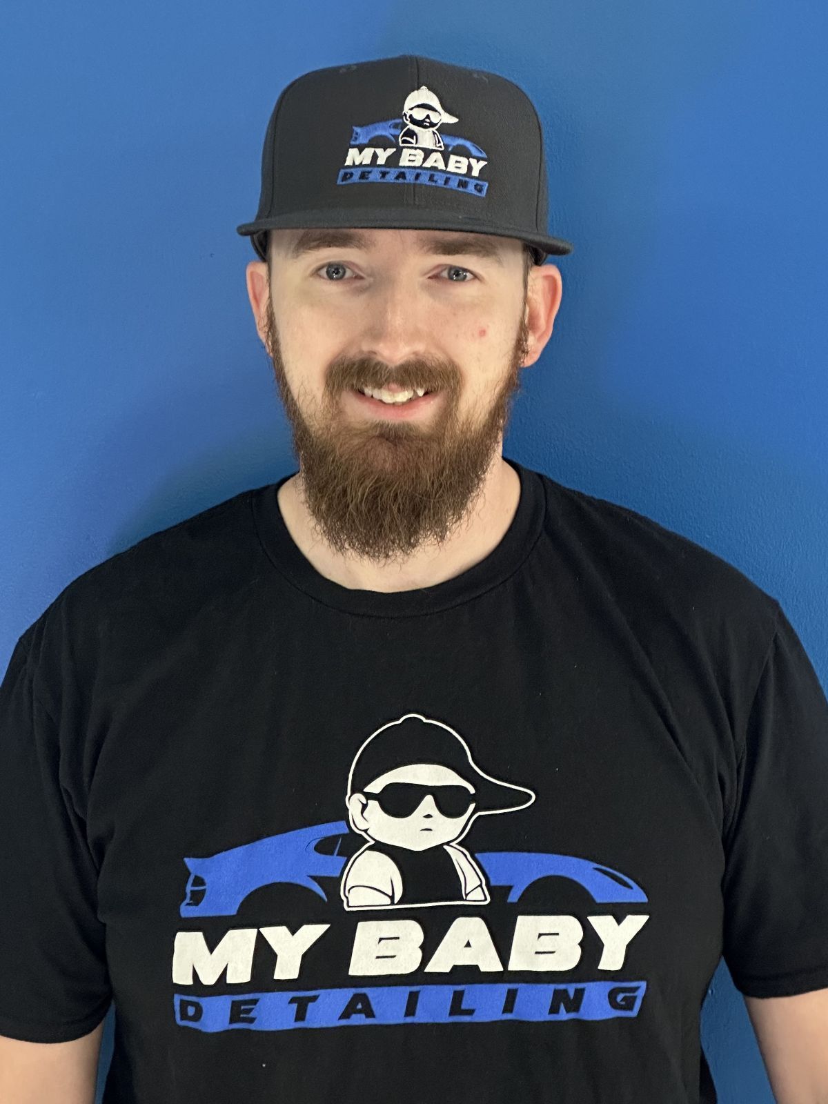 A person smiling against a bright blue background, wearing a black and blue collared shirt with a logo on the chest.