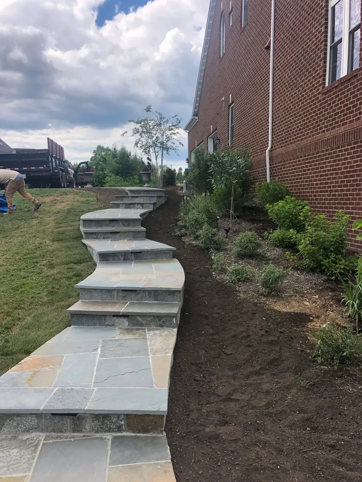 Stone steps curving up a grassy slope beside a brick house and landscaped side yard