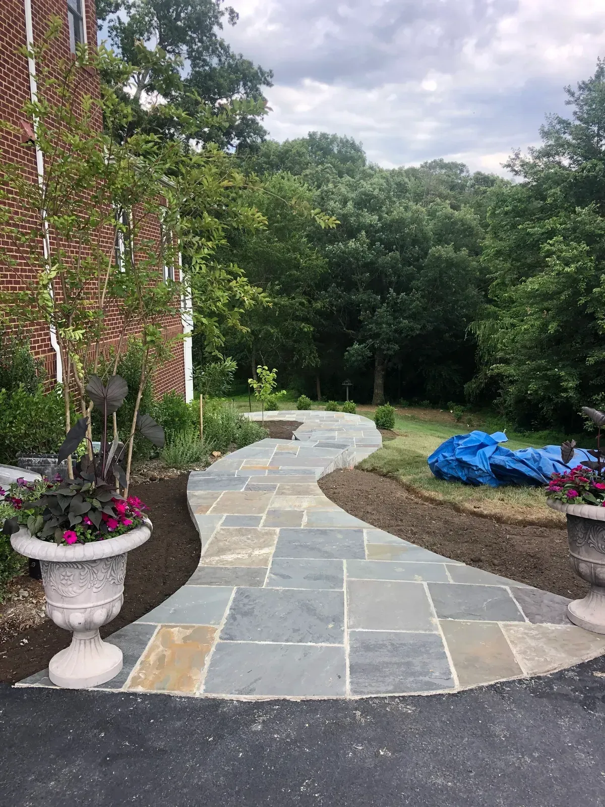 Curved stone garden path beside a brick house, potted flowers, and trees under a cloudy sky.
