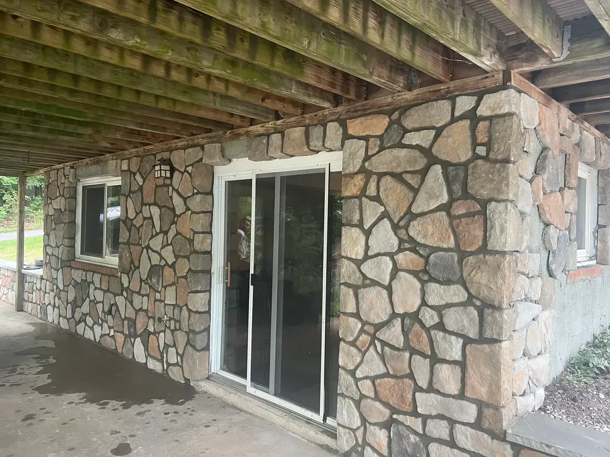 Stone-sided house patio with a sliding glass door and window under a wooden roof overhang
