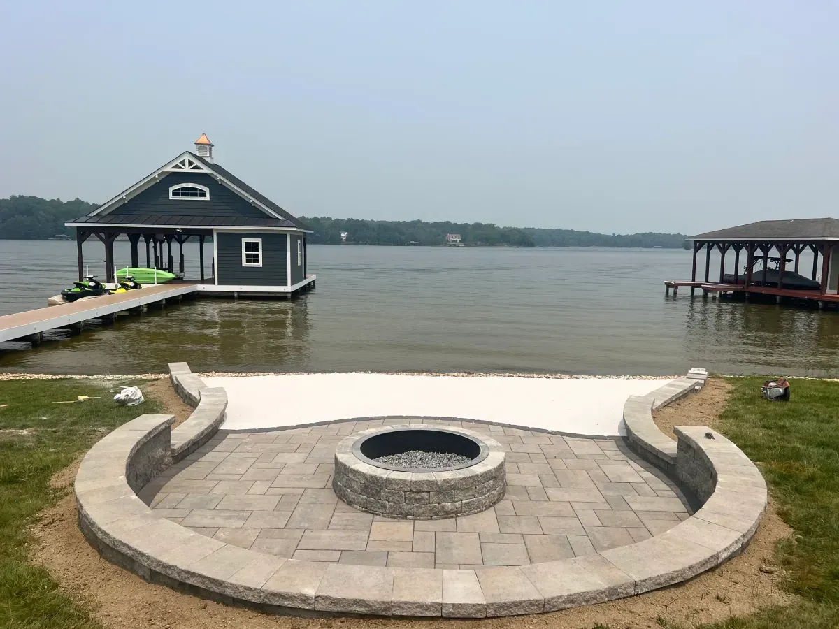 Lakeside fire pit patio with stone seating, docked boathouse, and calm water under an overcast sky