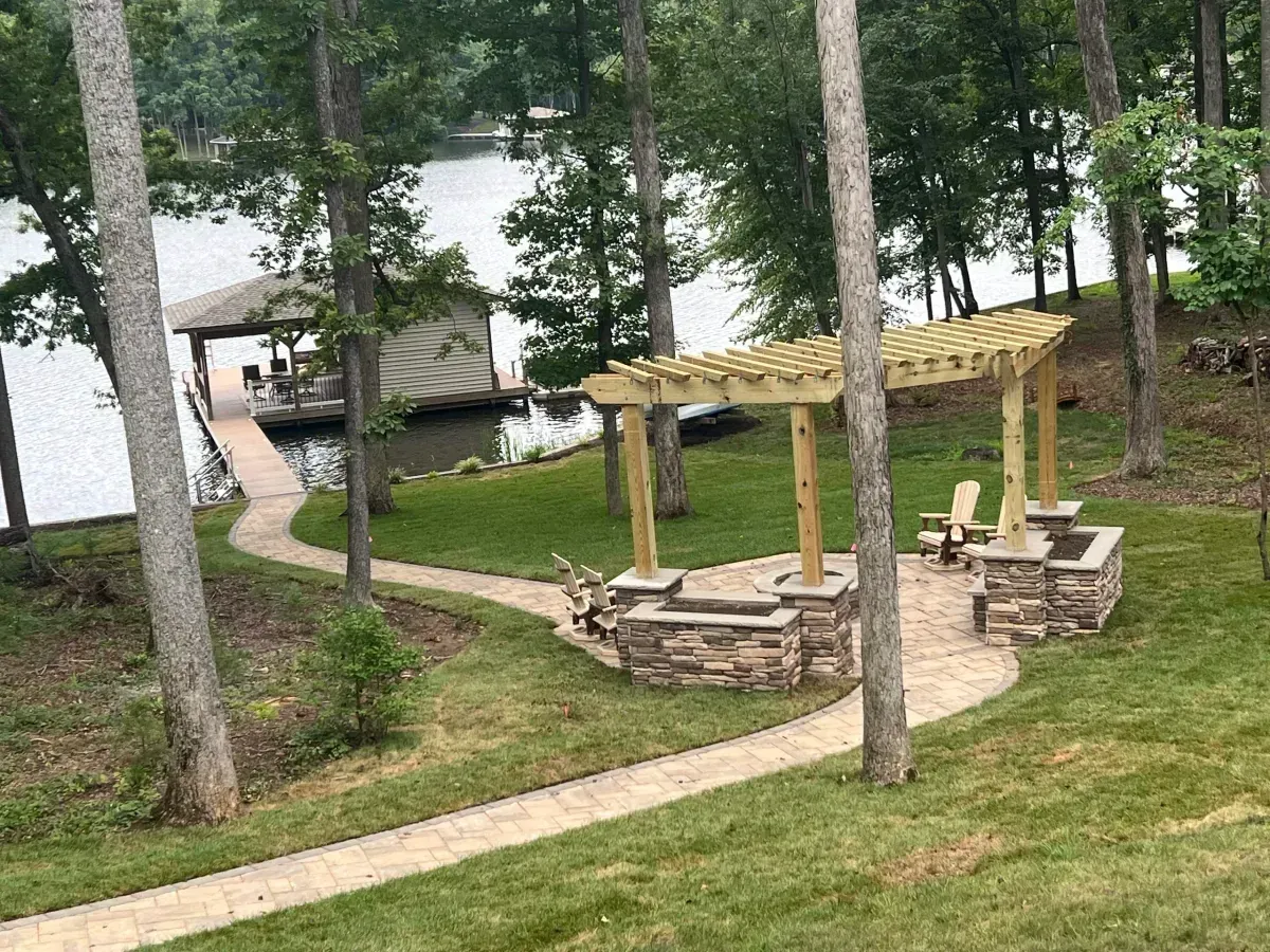 Stone patio and wooden pergola near a lakeside cabin among trees, with a winding path and picnic table on a grassy slope