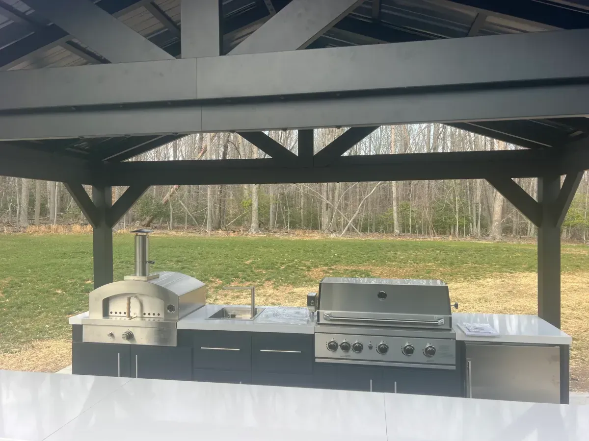 Covered outdoor patio with a stainless grill and brick pizza oven overlooking a grassy field